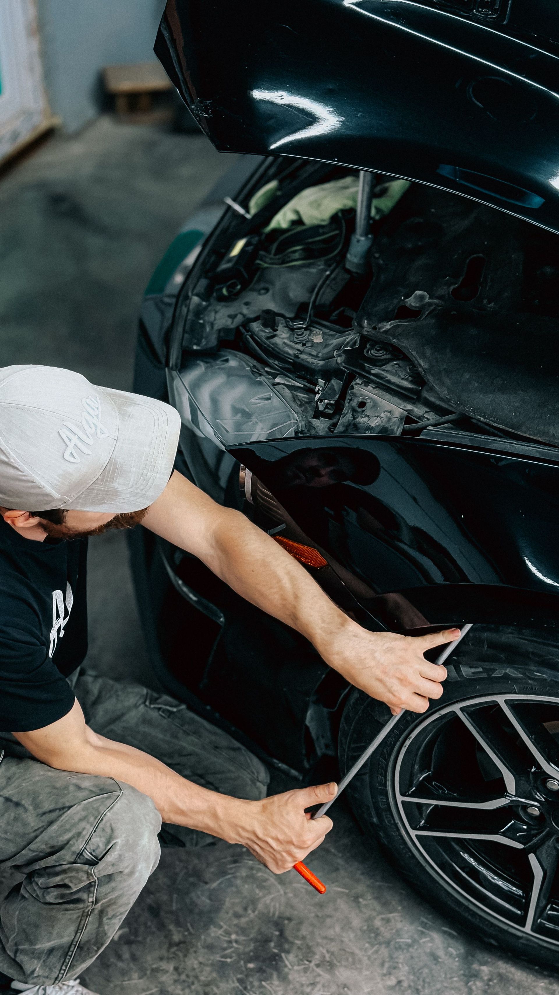 Mechanic inspecting a black car’s damaged front wheel and bumper in a garage