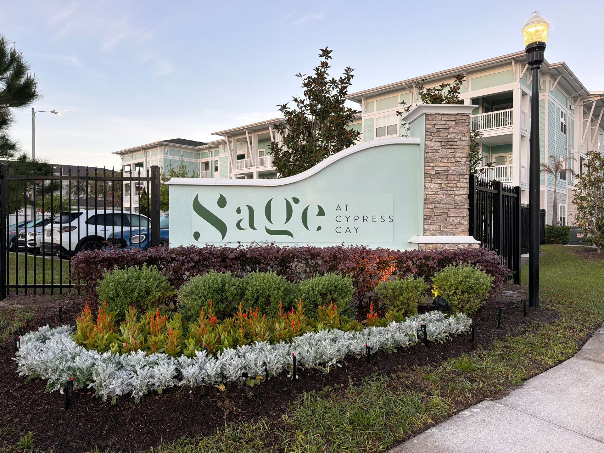 Apartment community entrance sign with landscaped flowers and black iron fence at dusk