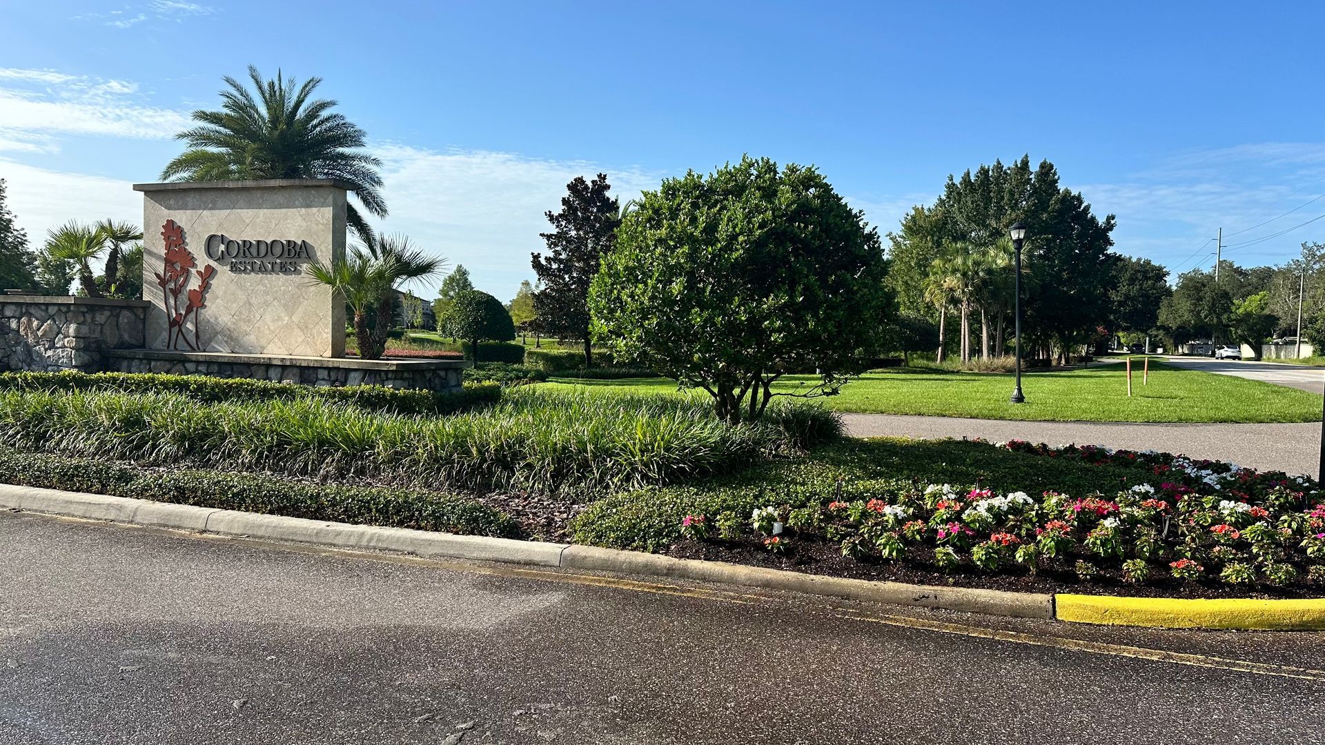 Landscape entrance sign beside a flower bed and manicured trees under a sunny blue sky