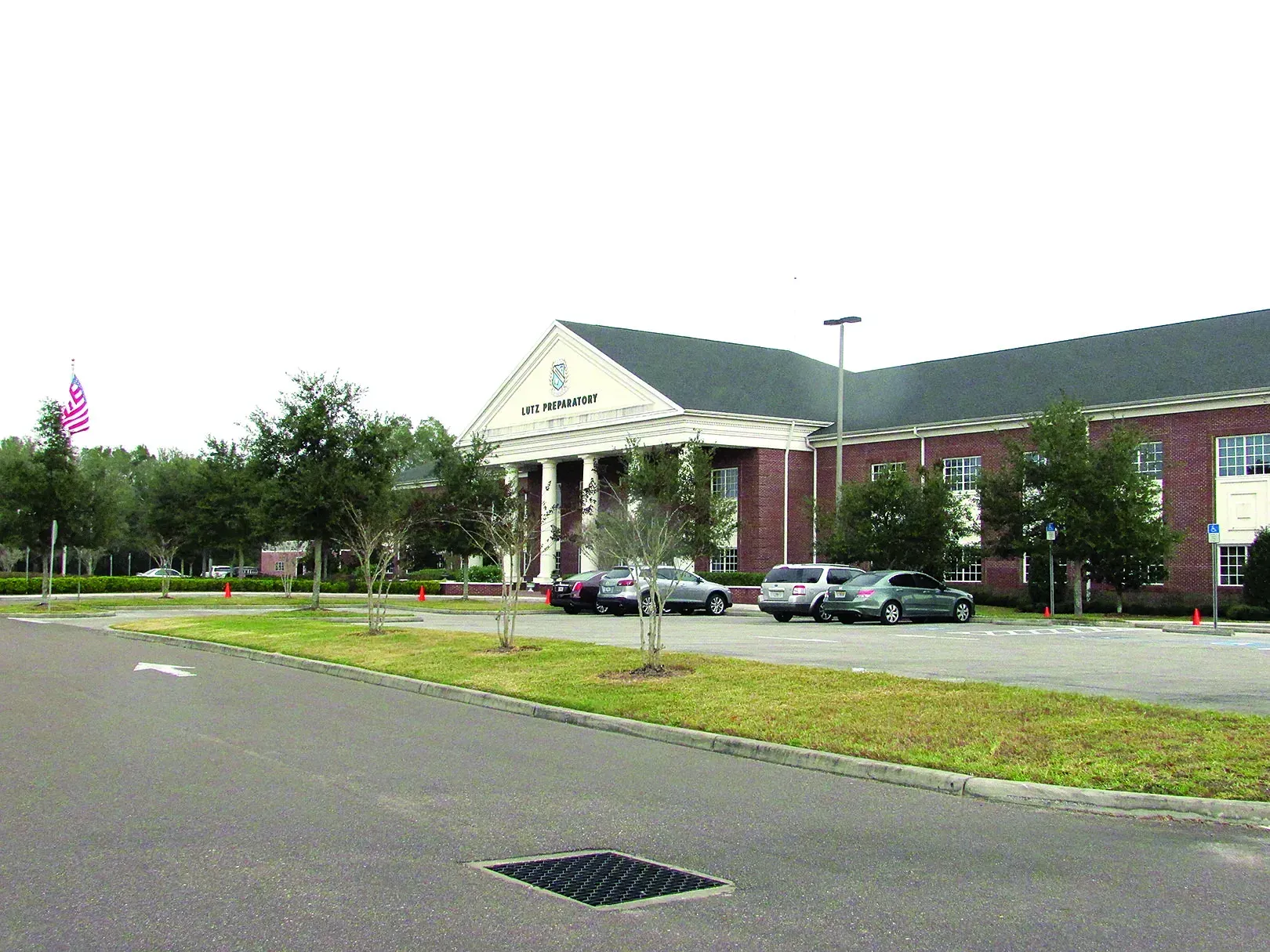 Parking lot and red-brick building with white columns, trees, and a road in the foreground