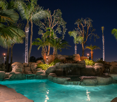Tropical resort pool at night with palm trees, glowing lights, and rock waterfalls