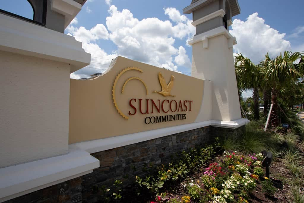 Suncoast Community entrance sign on a stucco wall with flowers and palm trees under a blue sky