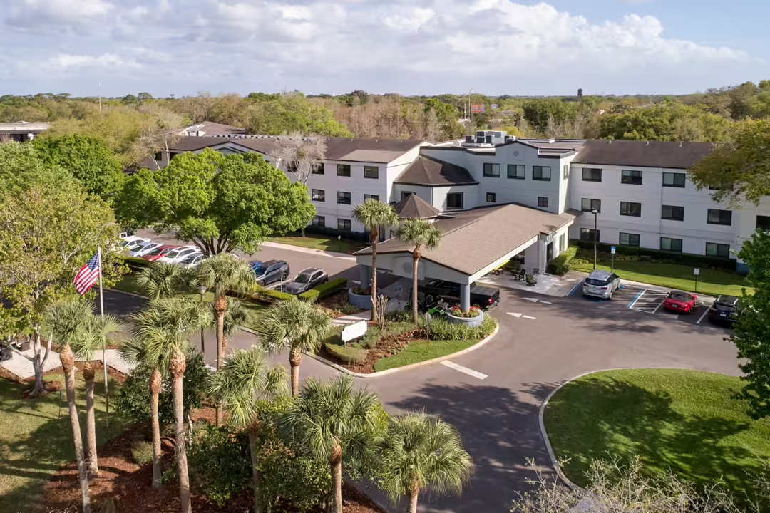 Aerial view of a hotel courtyard with palm trees, parking lot, and beige buildings under a blue sky