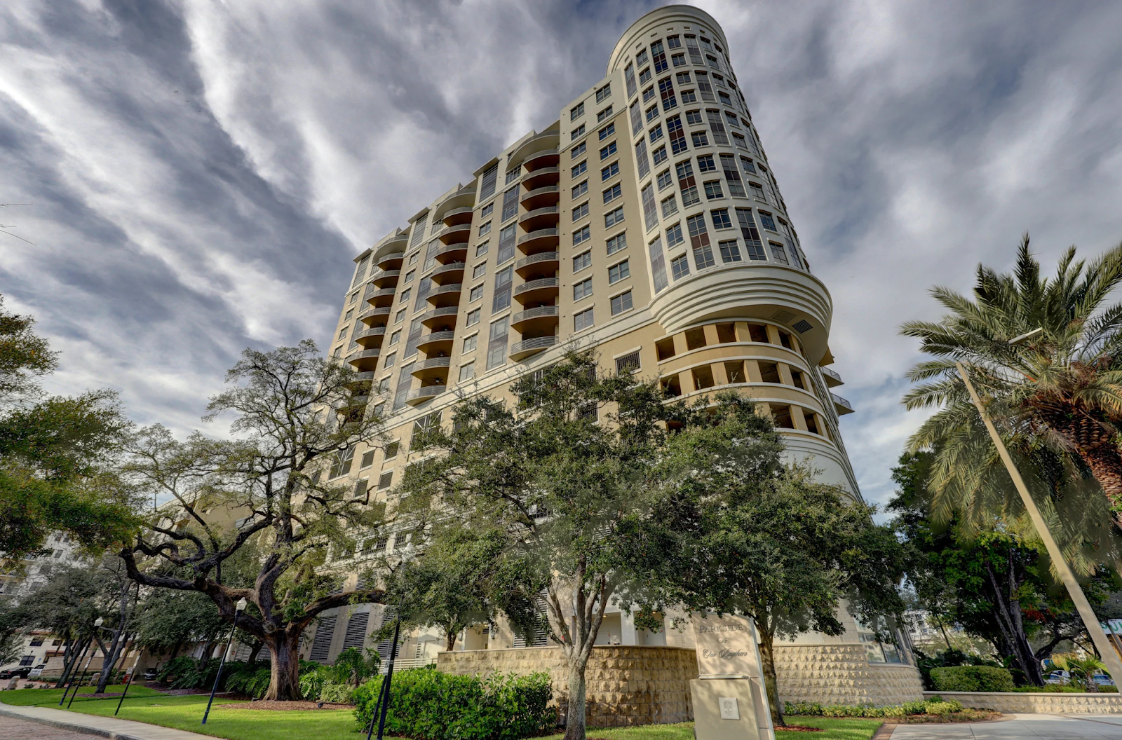 Tall beige high-rise building seen from below, framed by trees under a cloudy sky.