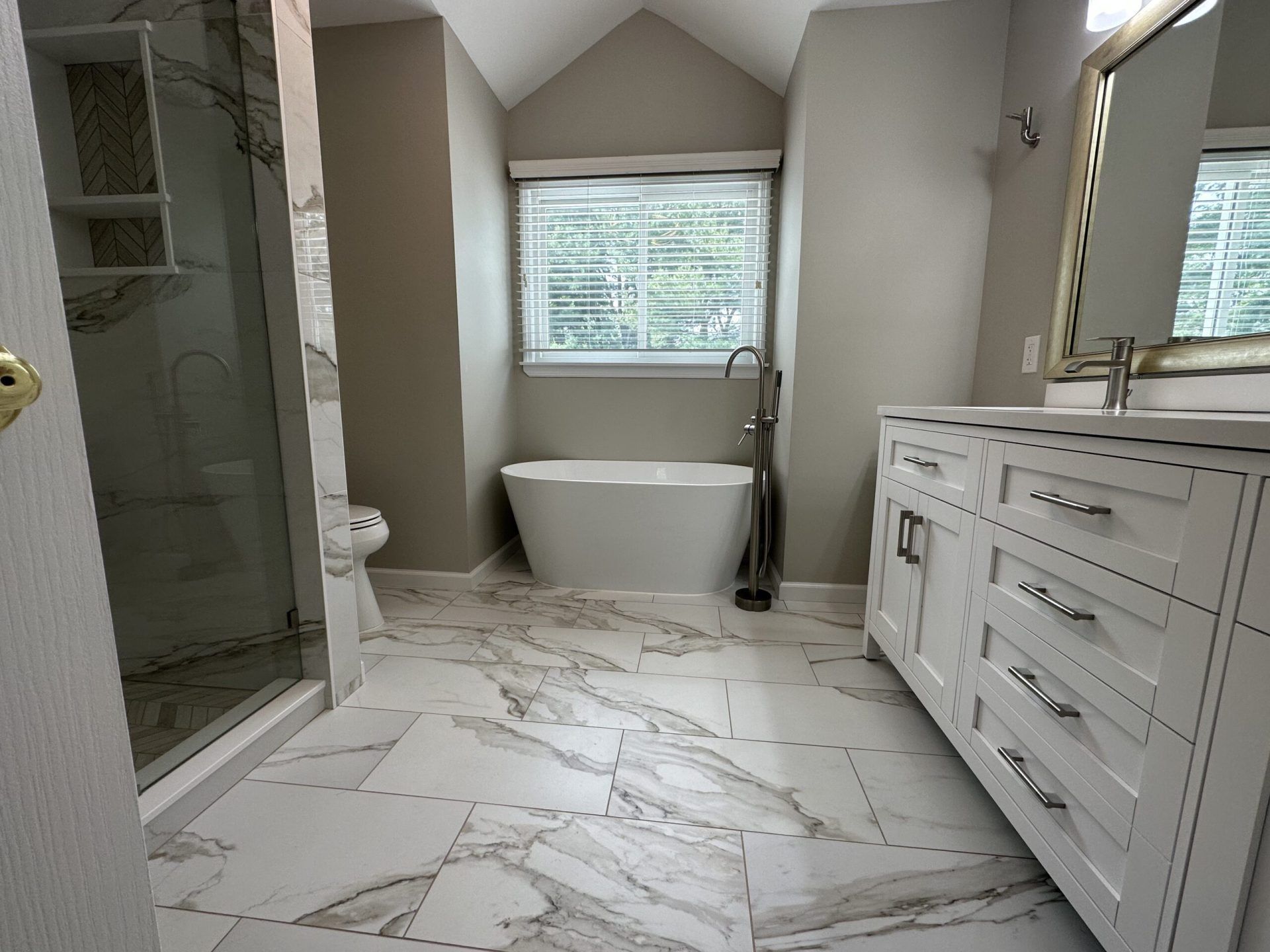 Bright marble bathroom with freestanding tub, glass shower, double vanity, and window