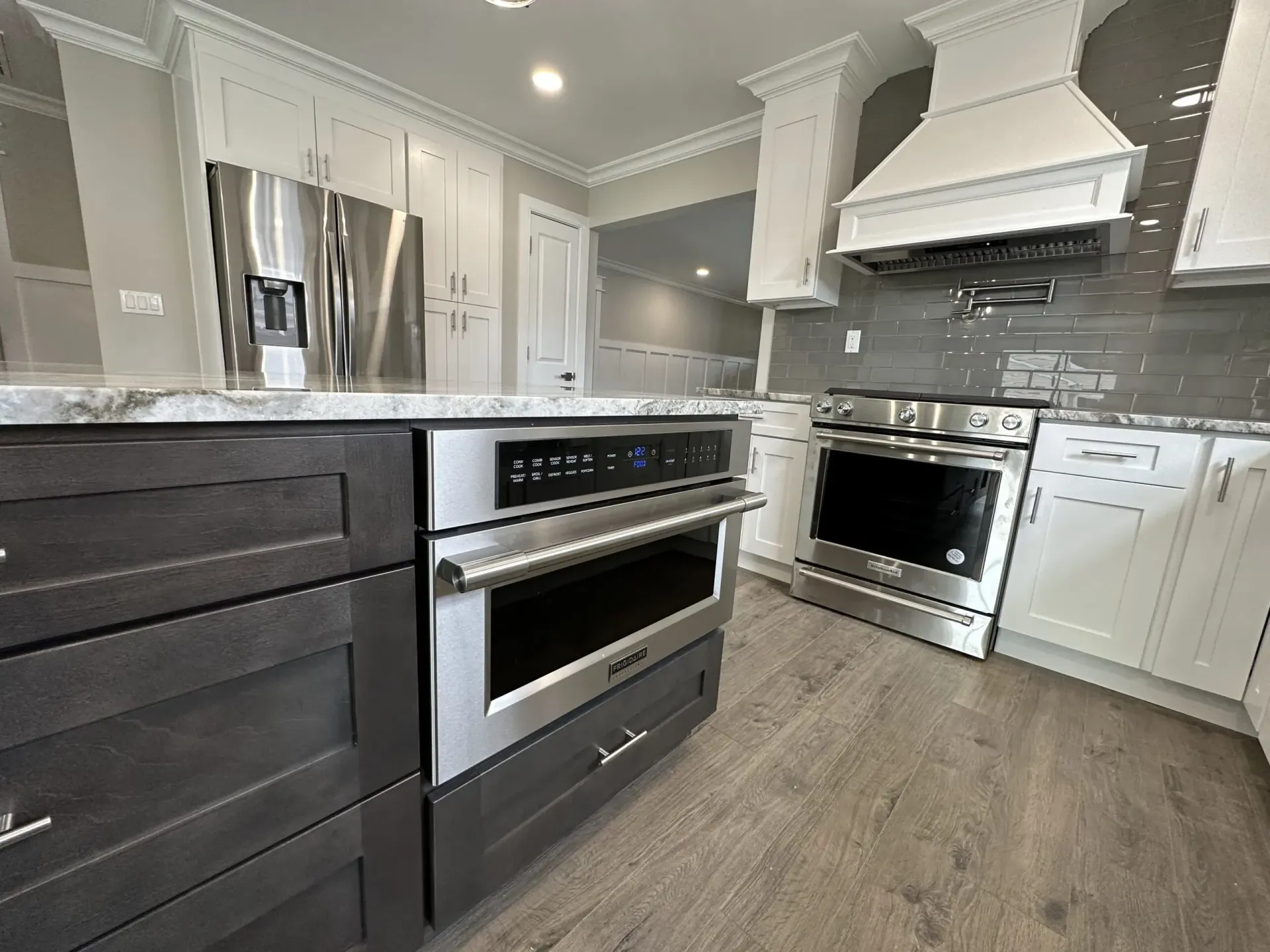 Modern white kitchen with stainless steel oven, range hood, and dark lower cabinets, shown from the island.