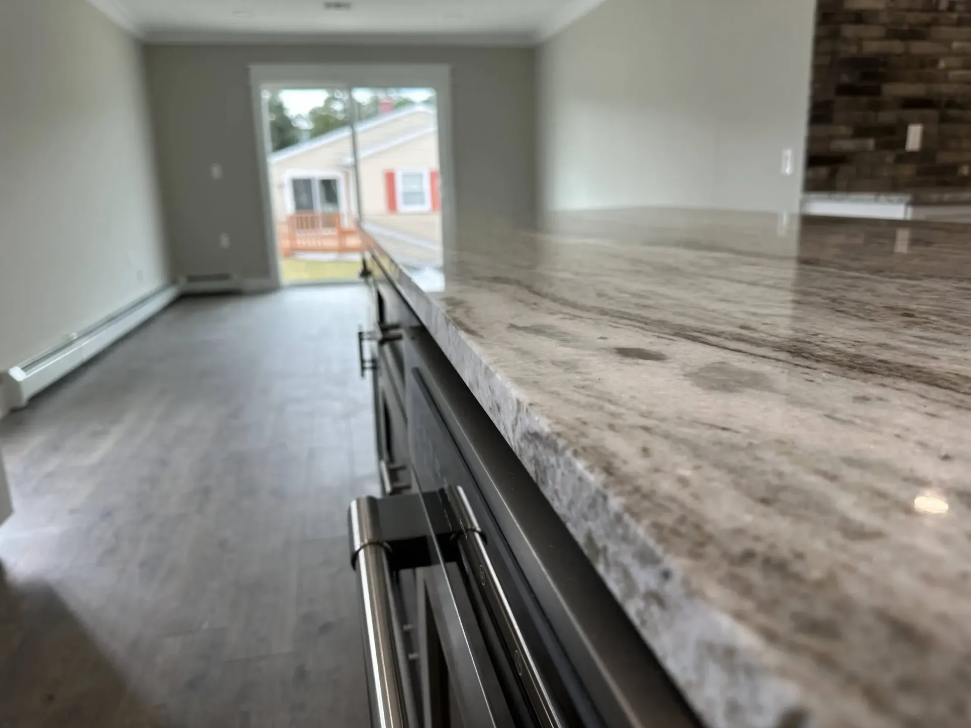 Empty modern kitchen with gray cabinets and a marble island, looking toward a bright window and backyard.