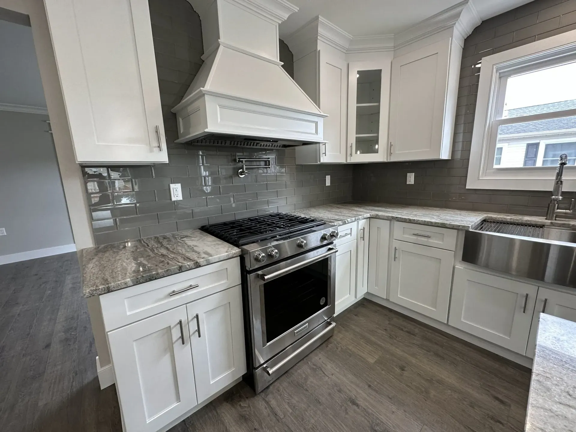 Modern white kitchen with granite counters, stainless steel stove, gray tile backsplash, and large window