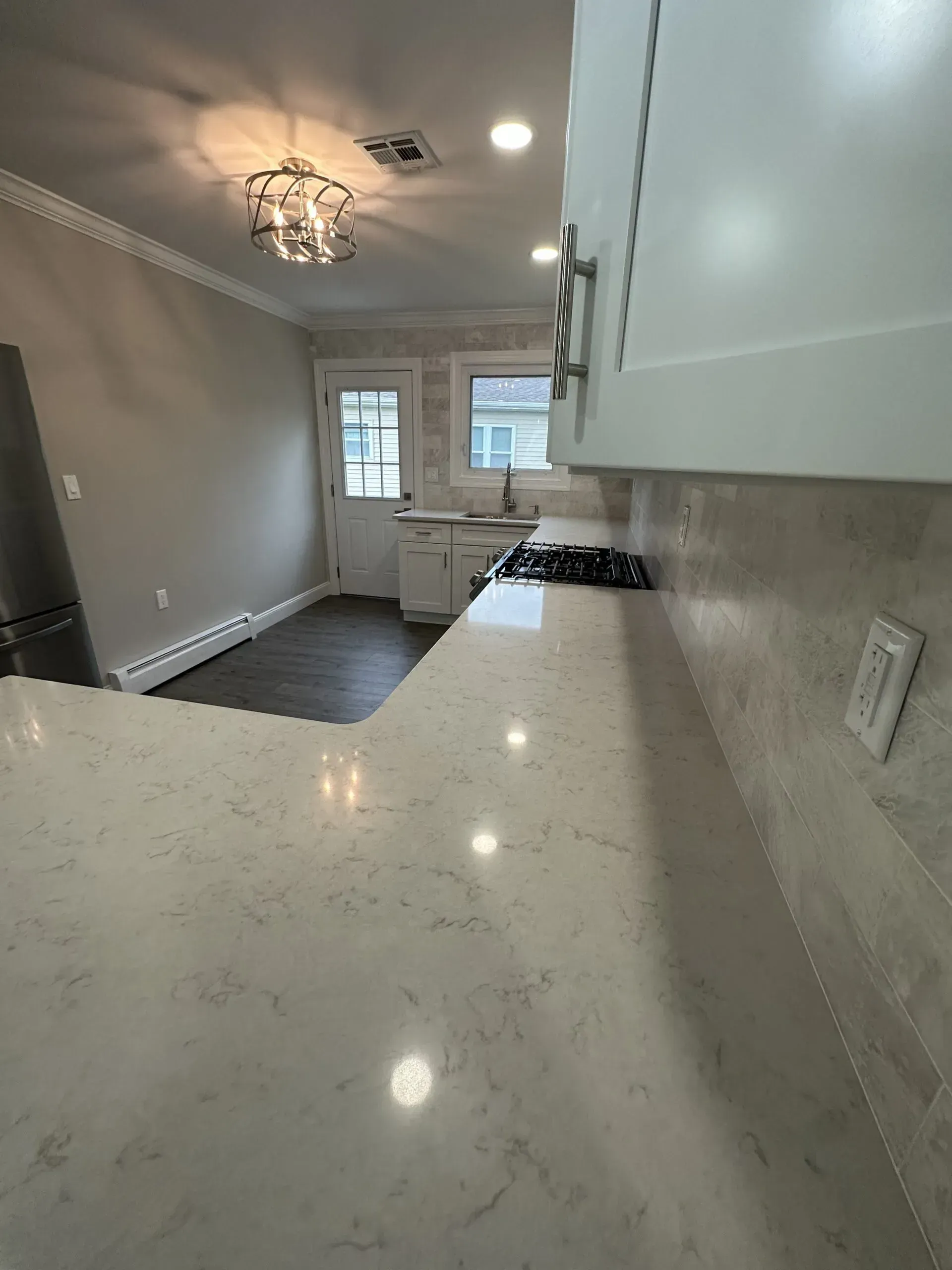 Empty modern kitchen with white countertops, gray walls, tile floor, and pendant light near a back door.