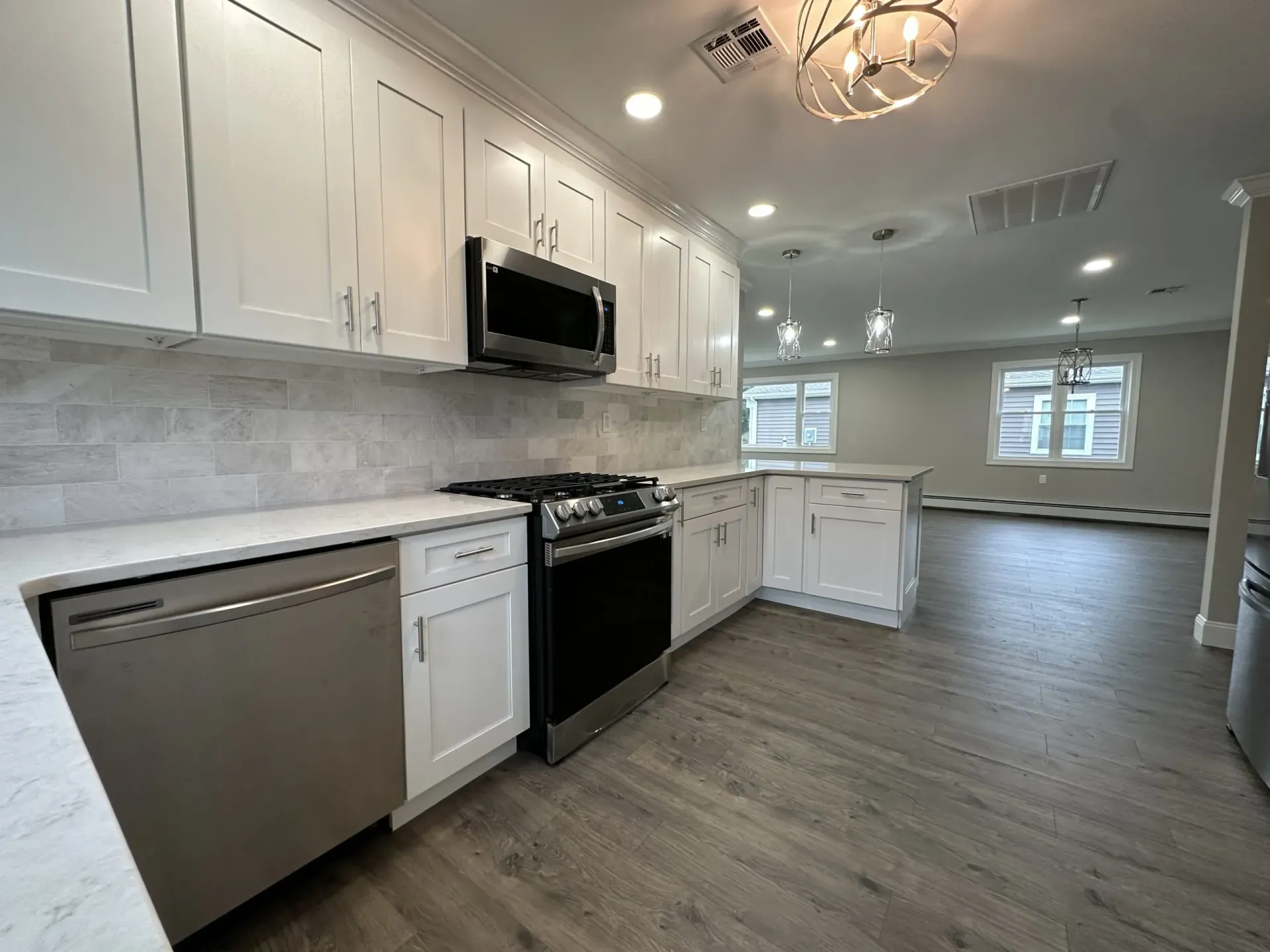 Bright modern kitchen with white cabinets, stainless appliances, and gray wood floors opening to a living area