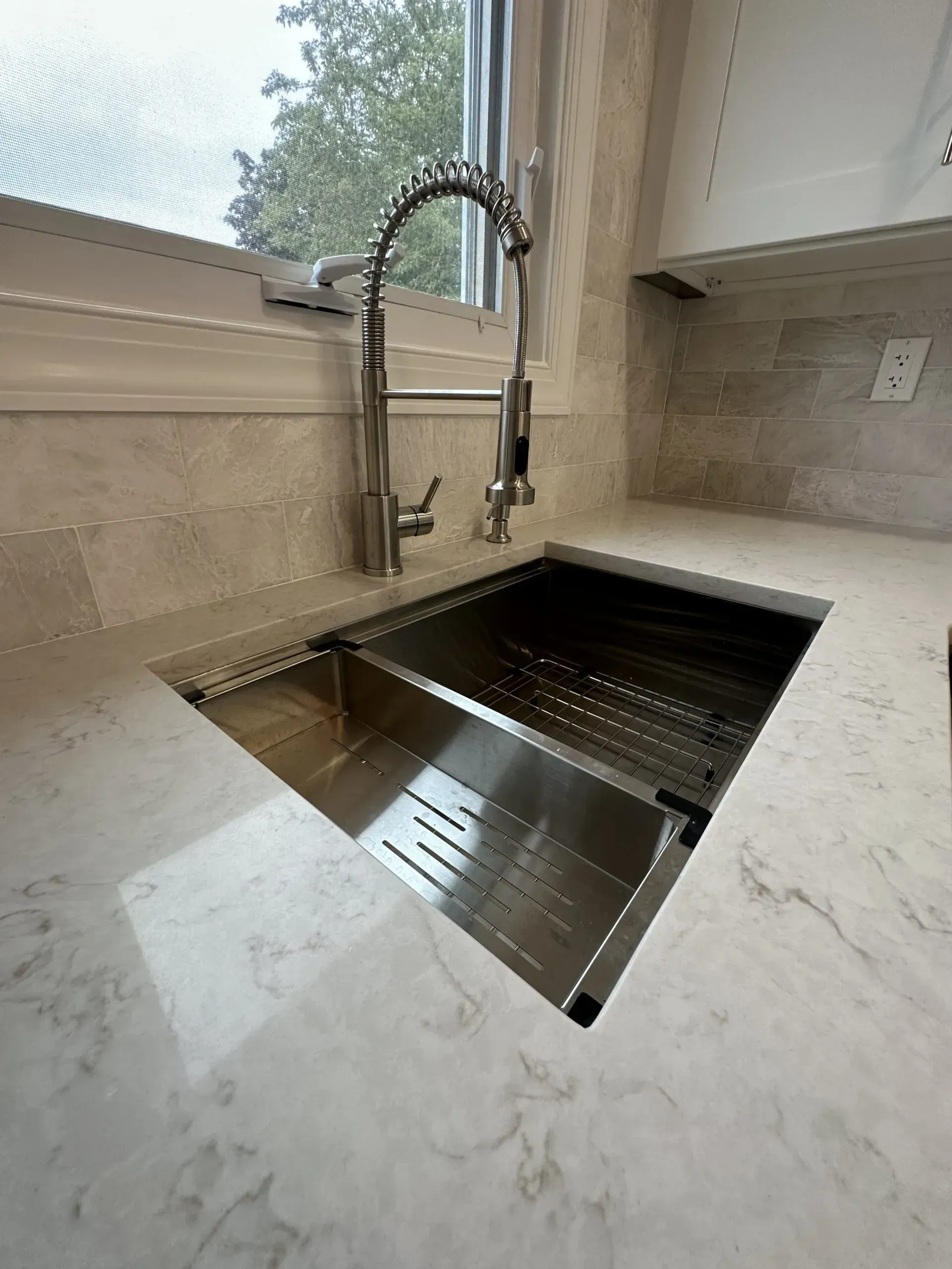 Modern kitchen sink with gooseneck faucet beside a window, set in a light marble countertop.