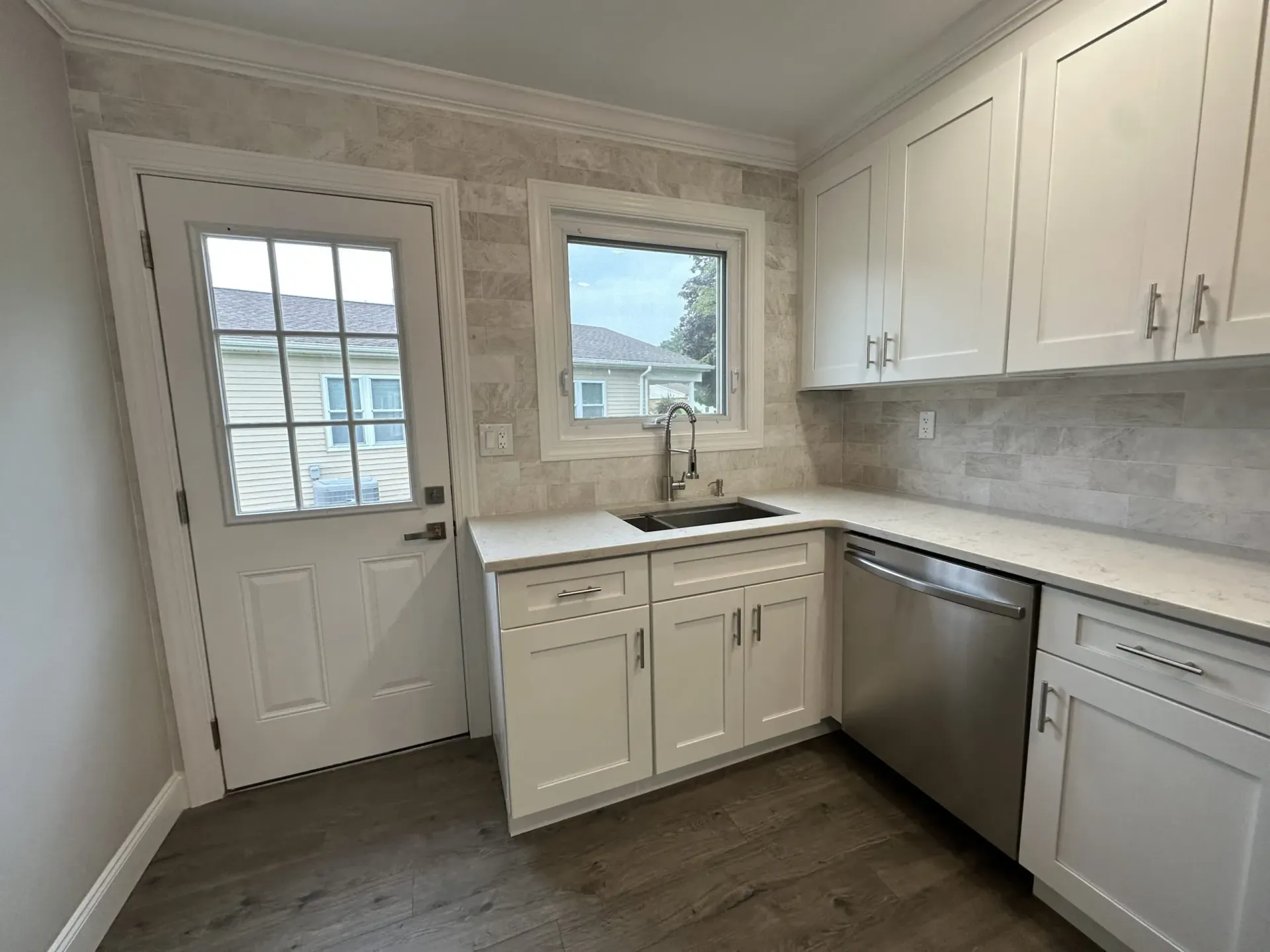 Bright white kitchen with cabinets, stainless dishwasher, sink, and door to outside.