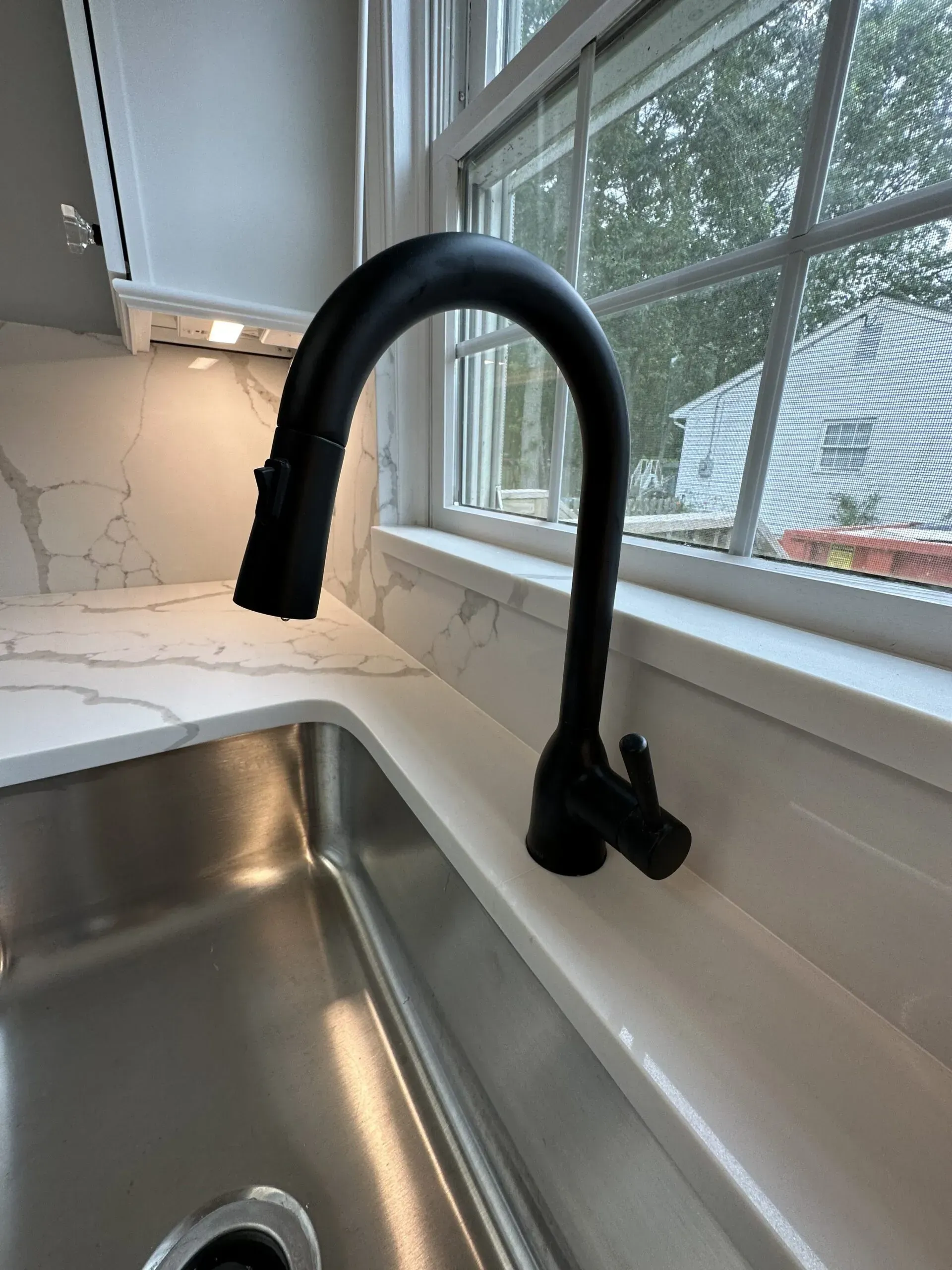 Black kitchen faucet over stainless sink beside a window and white countertop.