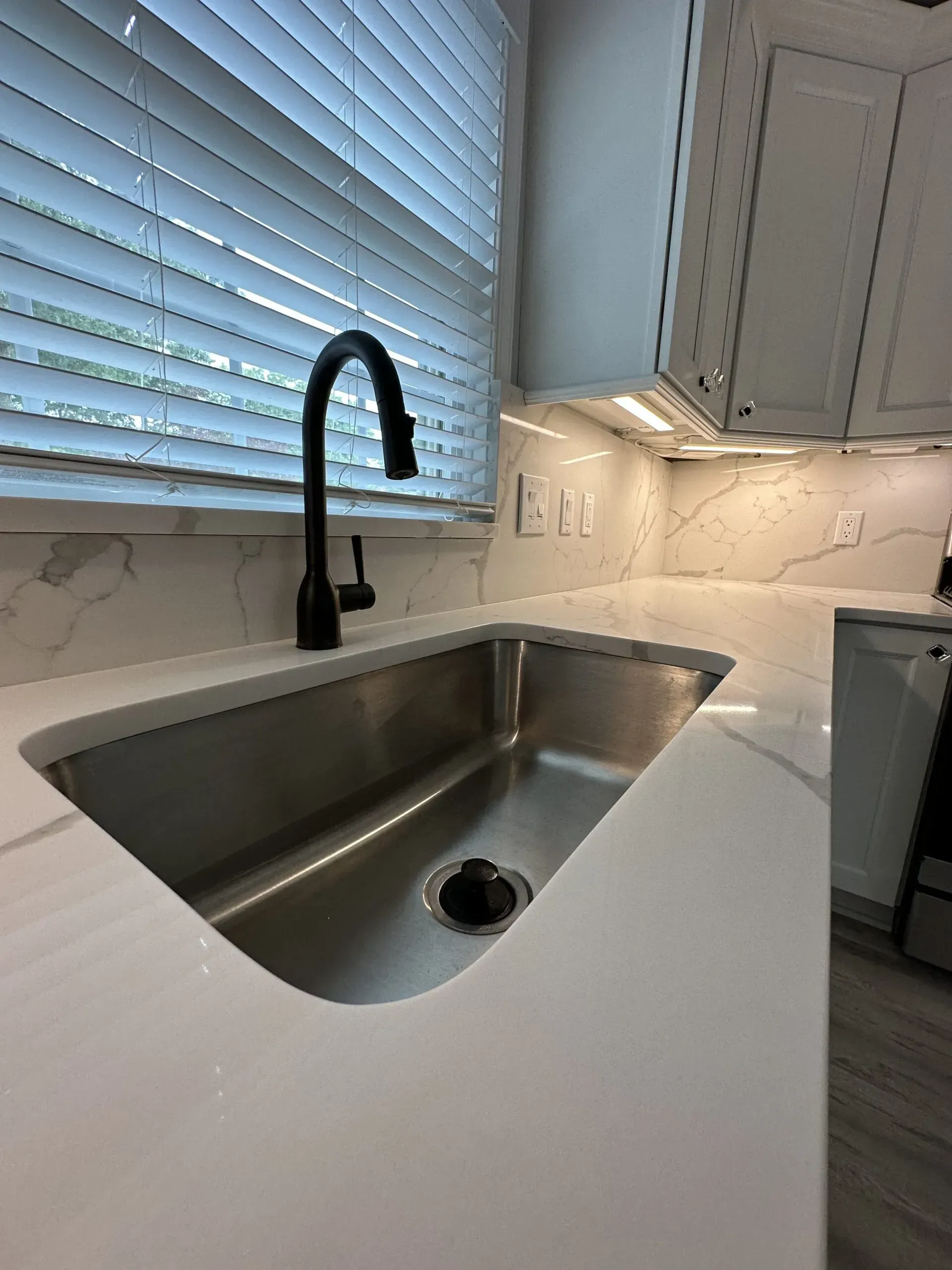 Modern kitchen sink with black faucet, white countertop, and window blinds behind it