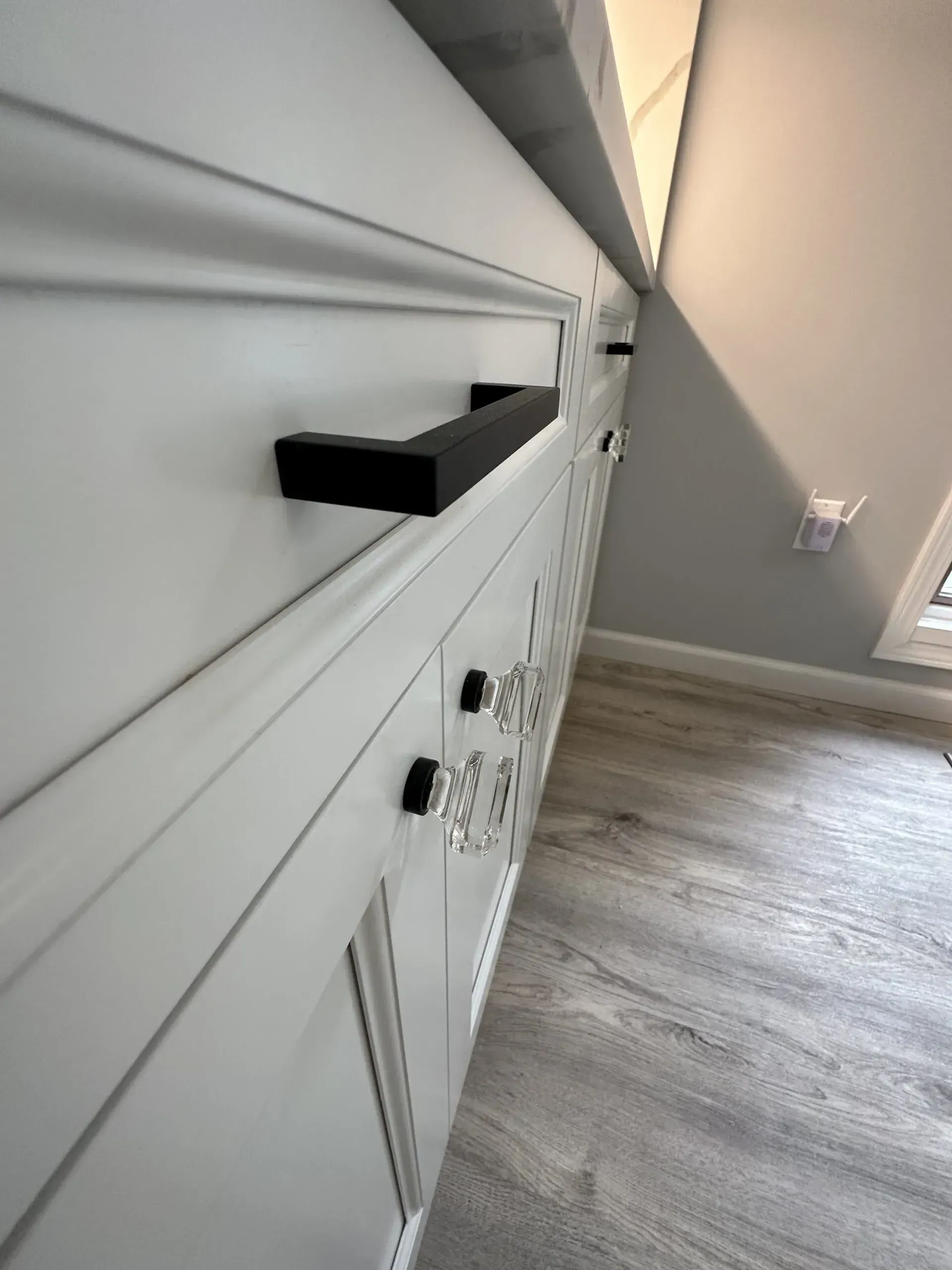 White built-in cabinetry with black handles in a bright hallway beside a gray wall and wood flooring