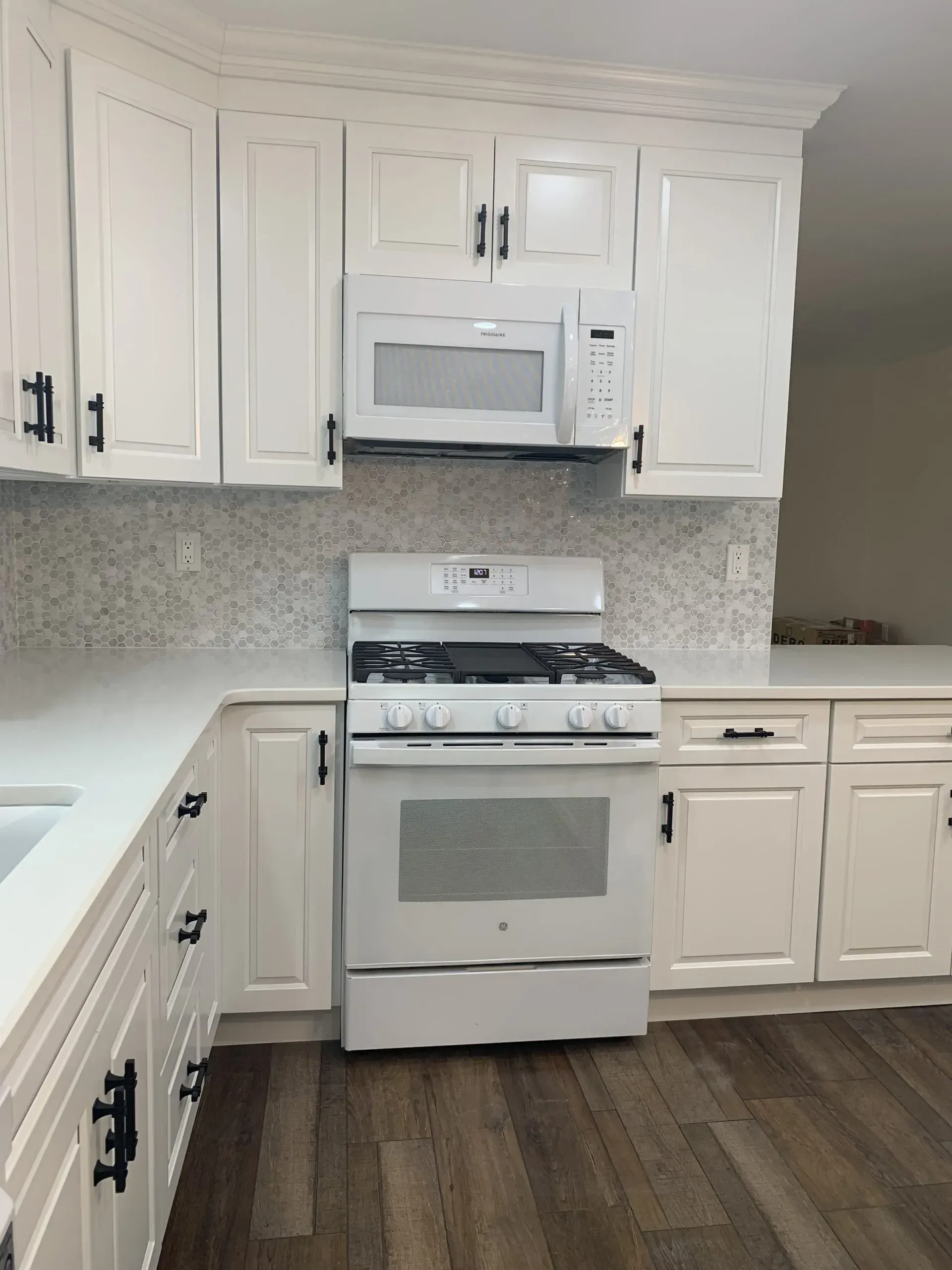 White kitchen with white cabinets, gas stove, microwave, sink, and dark wood flooring.