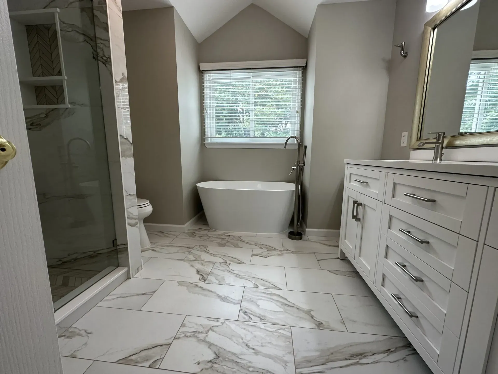 Modern bathroom with marble floor, freestanding tub, glass shower, and white vanity under a window