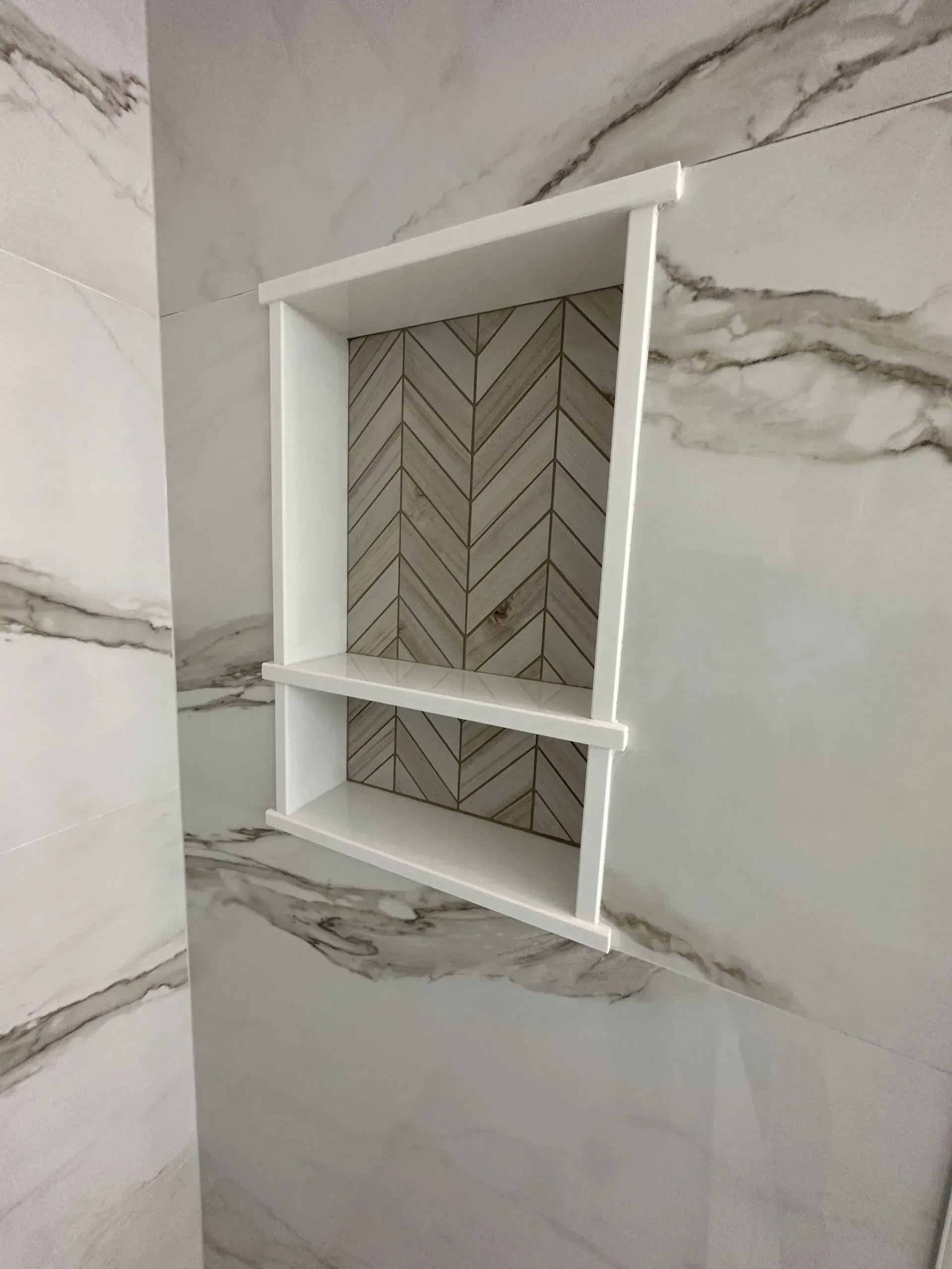 White recessed wall shelf in a marble-tiled shower with gray veining and a herringbone back panel