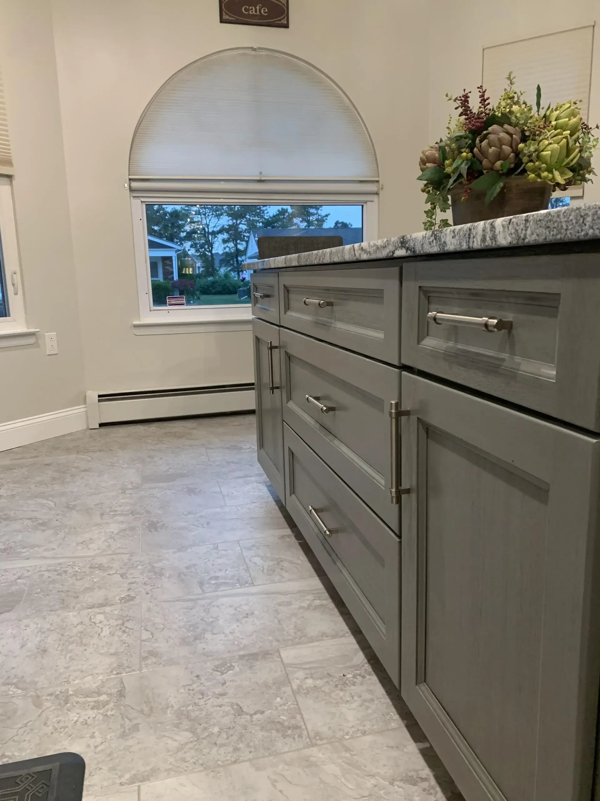 Gray kitchen island with drawers and a granite countertop, in a bright room with an arched window blind.