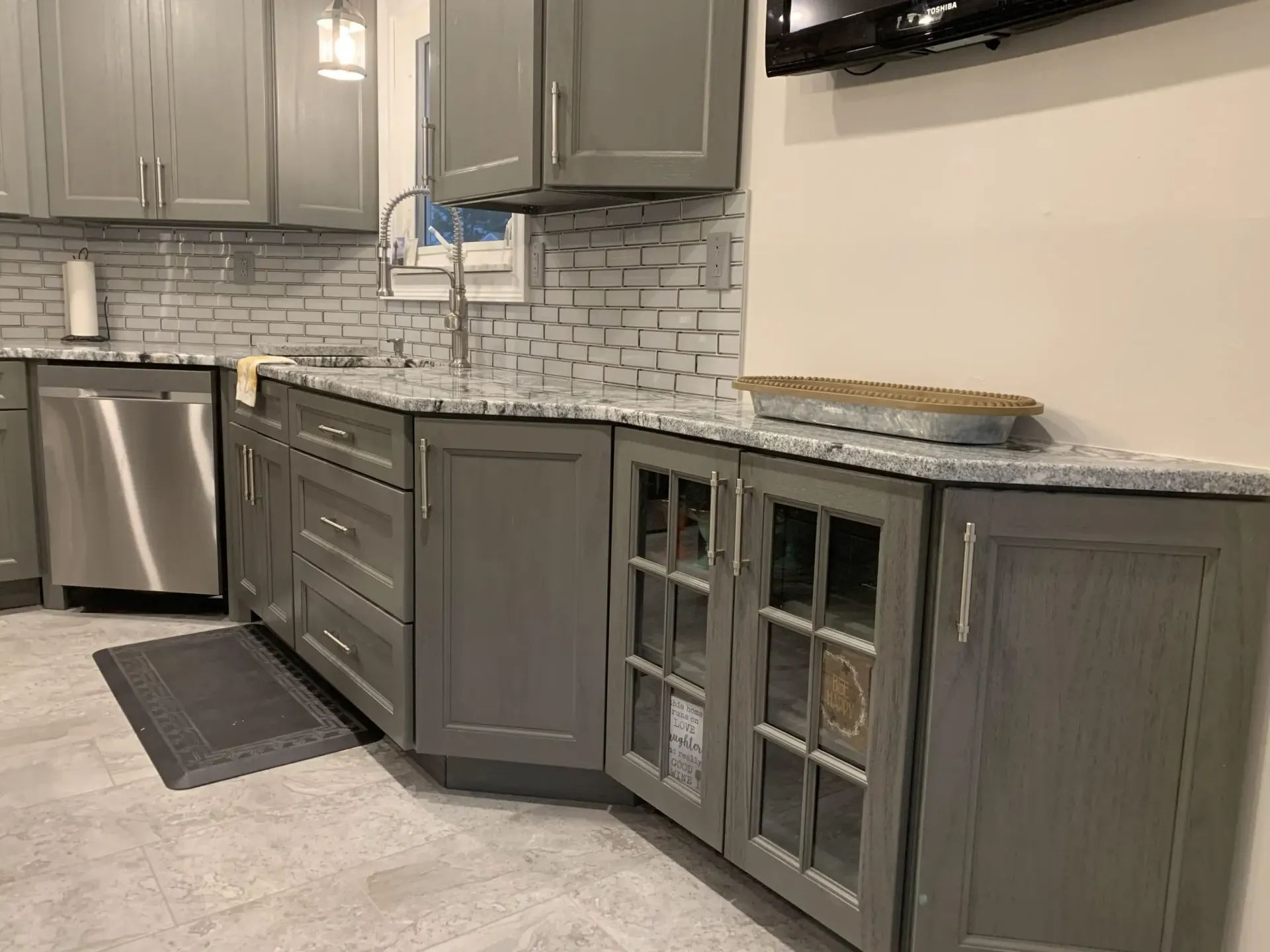 Modern kitchen with gray cabinets, stainless dishwasher, and glass-front lower cabinet beneath a marble-look countertop