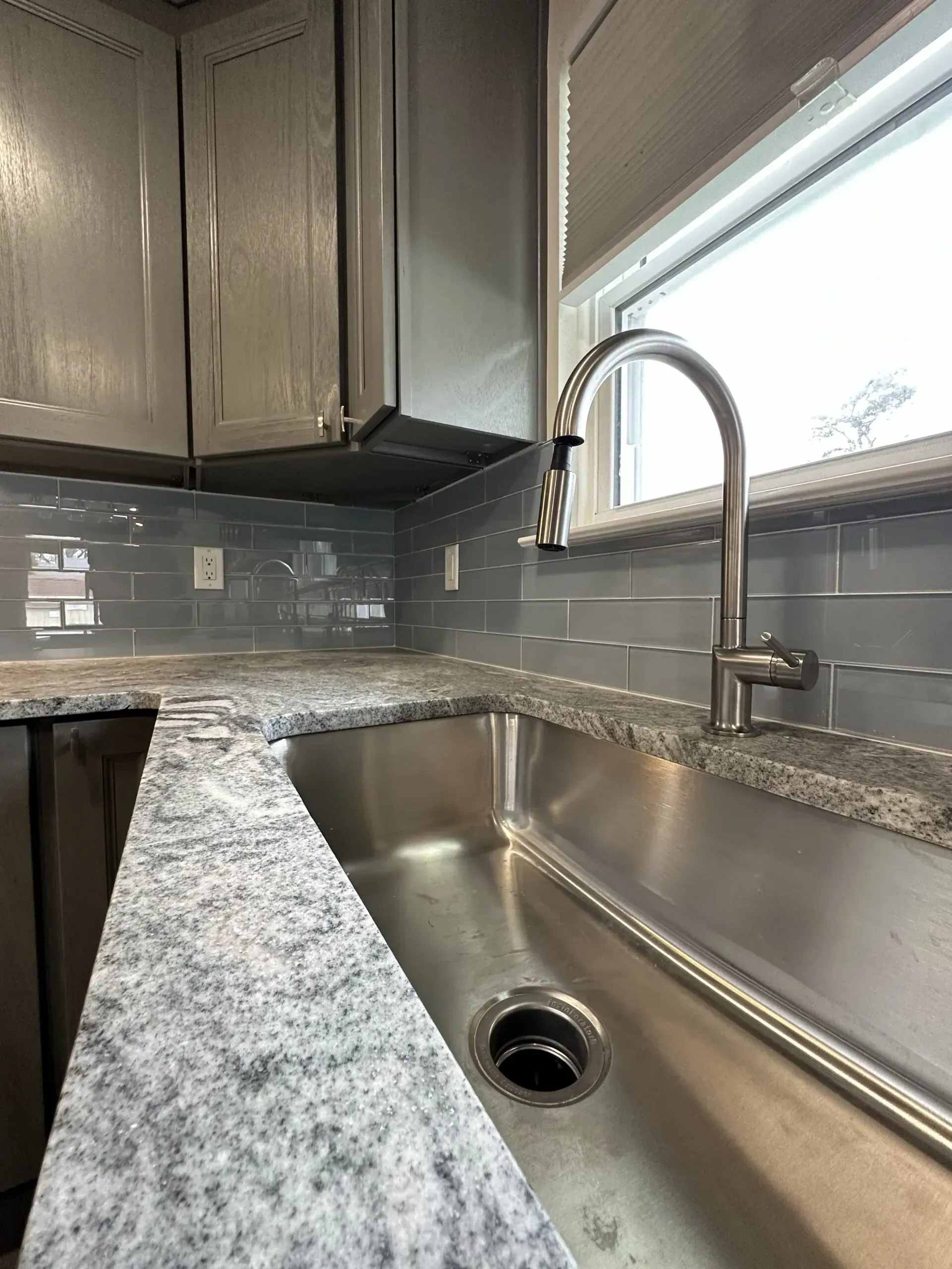 Modern kitchen sink with stainless steel basin, granite countertop, gray cabinets, and window above faucet