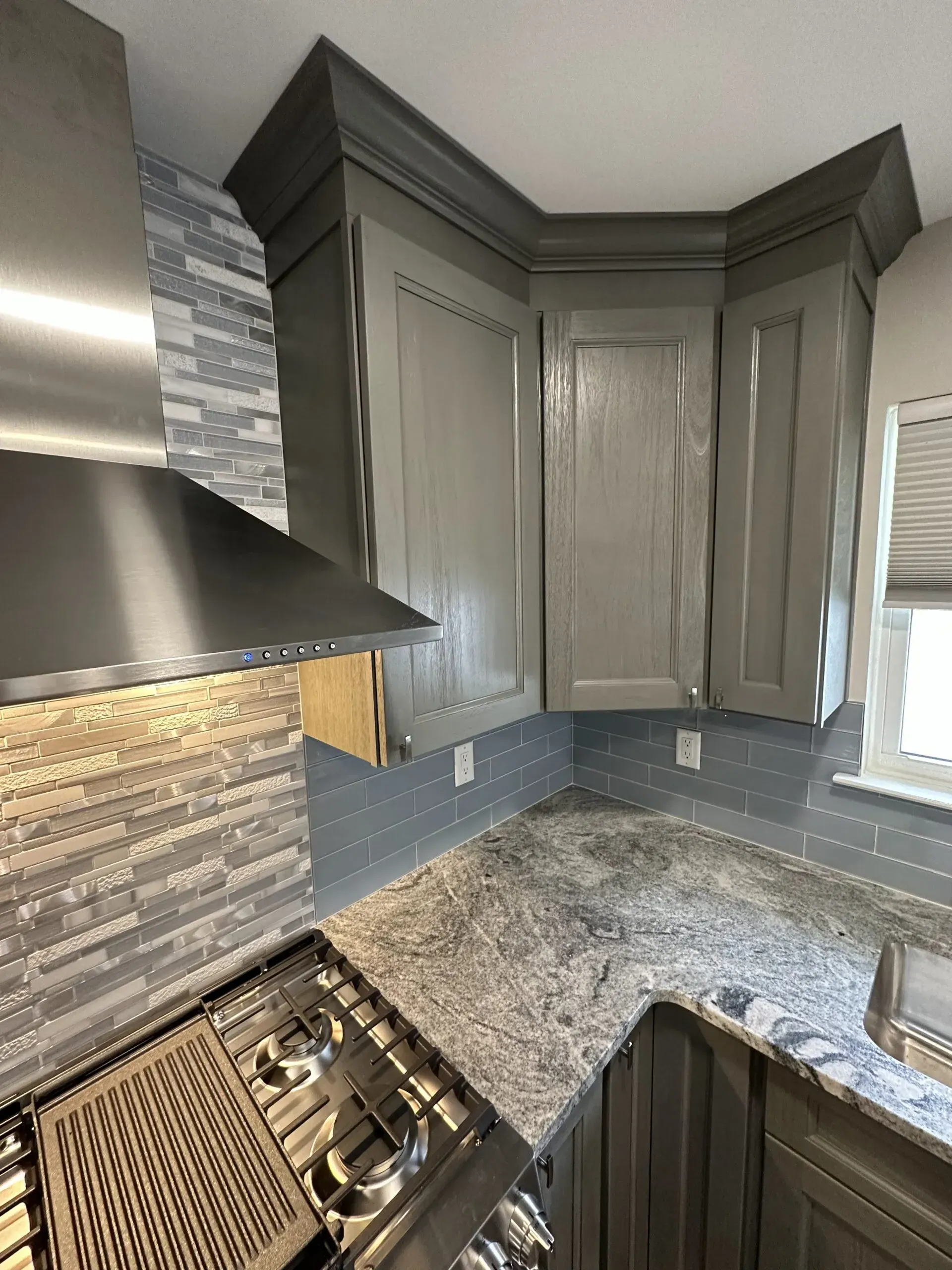 Modern kitchen corner with gray cabinets, granite countertops, stainless range hood, and gas cooktop