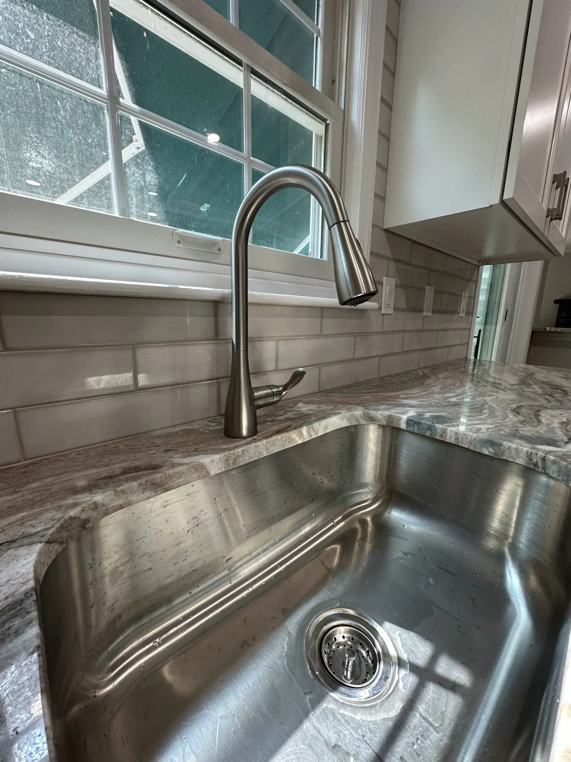 Stainless steel kitchen sink with chrome faucet beside a window and tiled backsplash.