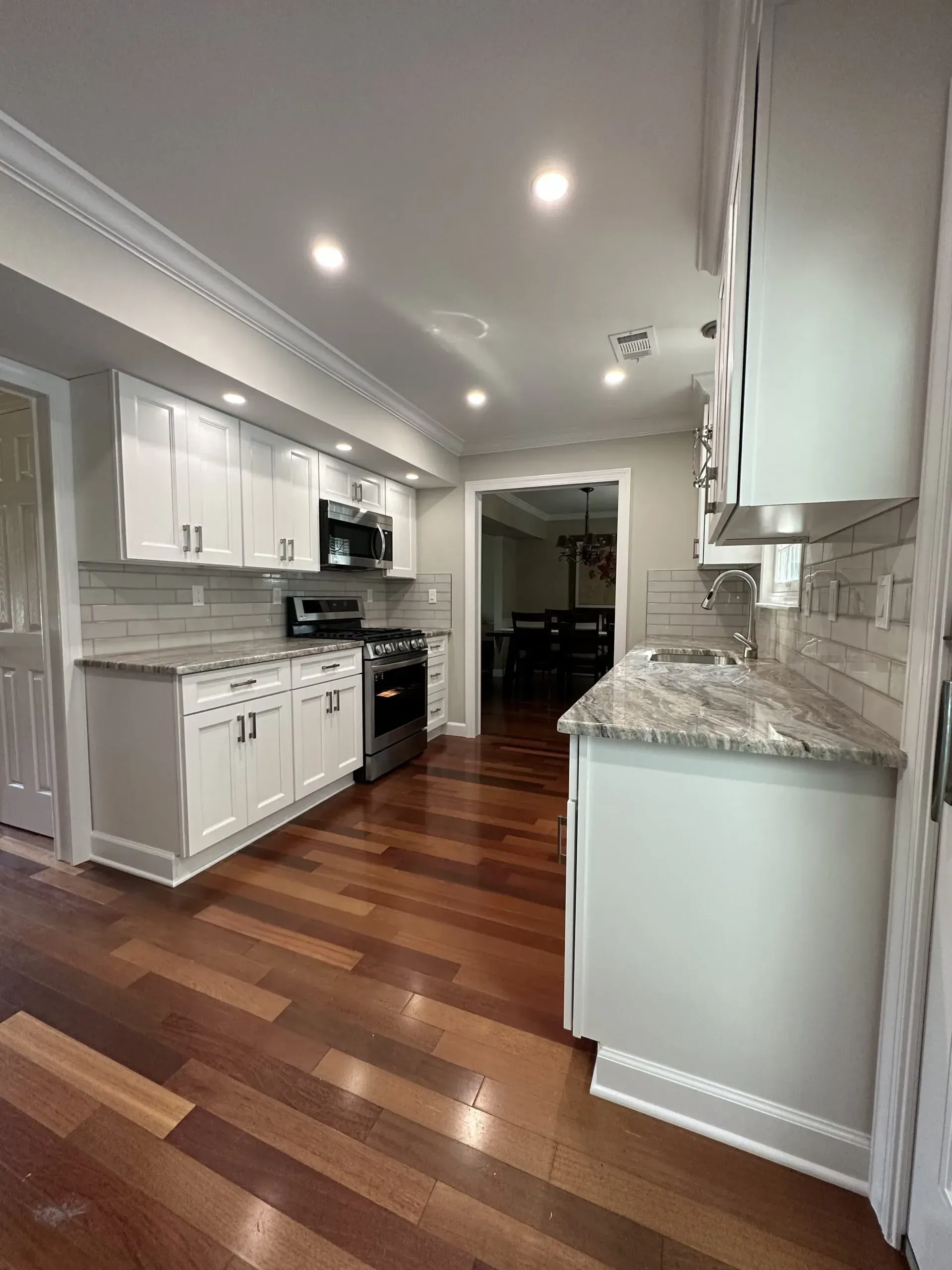 Bright white kitchen with granite counters, stainless appliances, and wood floors leading to a dining area