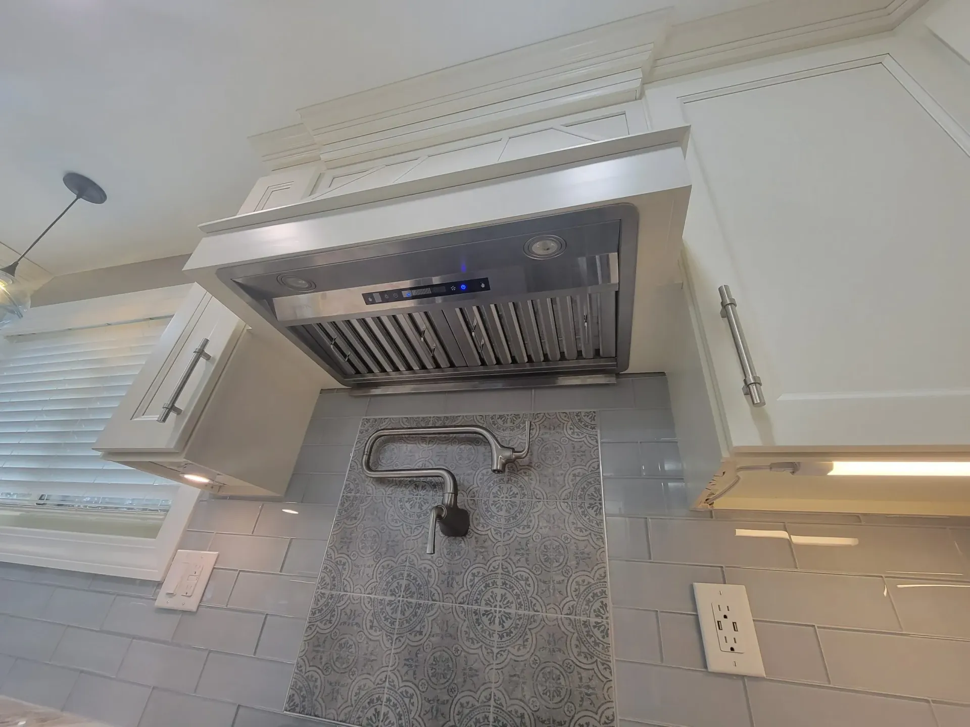 Modern kitchen sink under a stainless steel range hood with white cabinets and gray tile backsplash