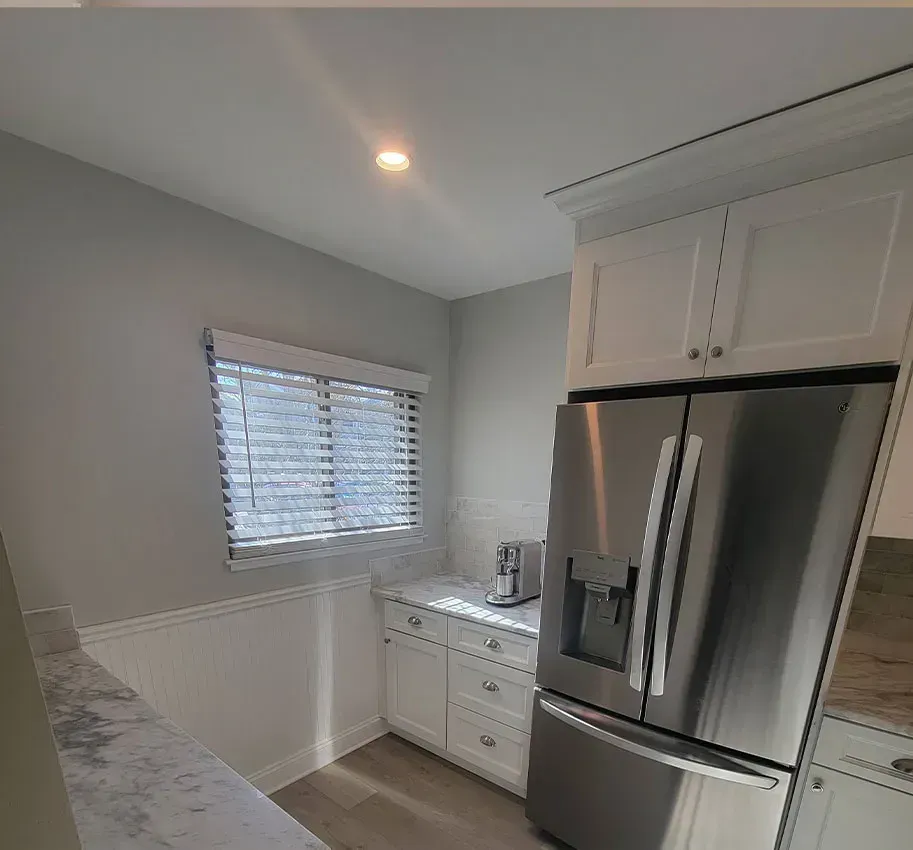 Bright white kitchen with stainless steel fridge, white cabinets, and a window with blinds