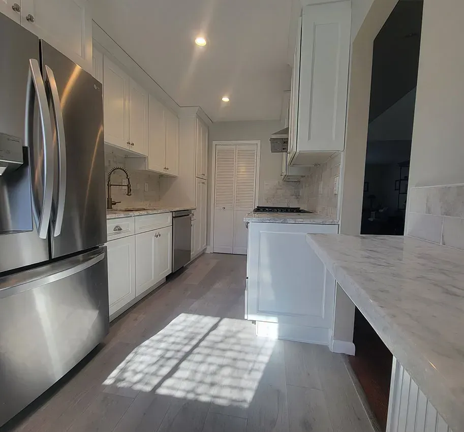 Bright galley kitchen with stainless steel fridge, white cabinets, and sunlight on gray flooring
