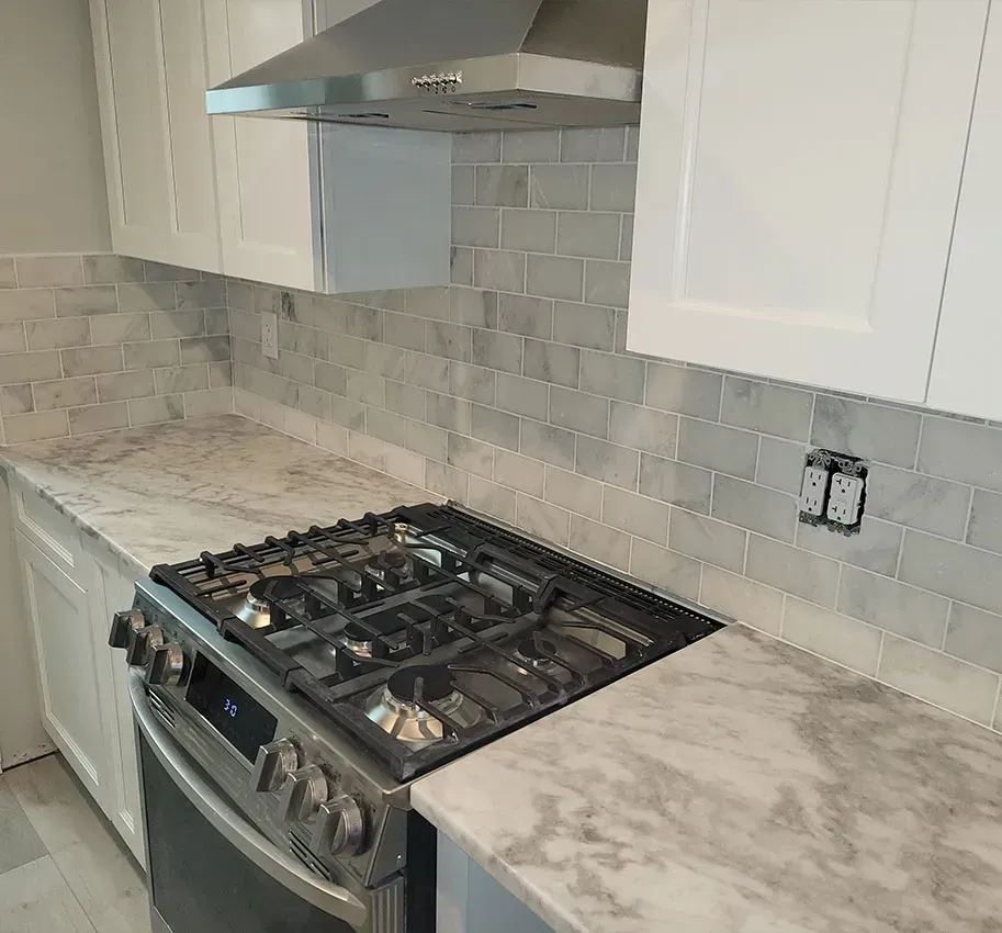 Modern kitchen with white cabinets, gray tile backsplash, stainless range hood, and gas stove beside marble-look counters