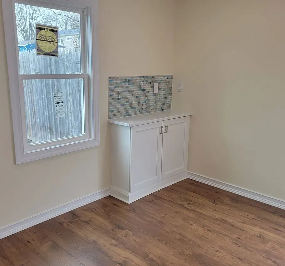 Empty room with wood floors, window, and small white cabinet with mosaic tile backsplash.