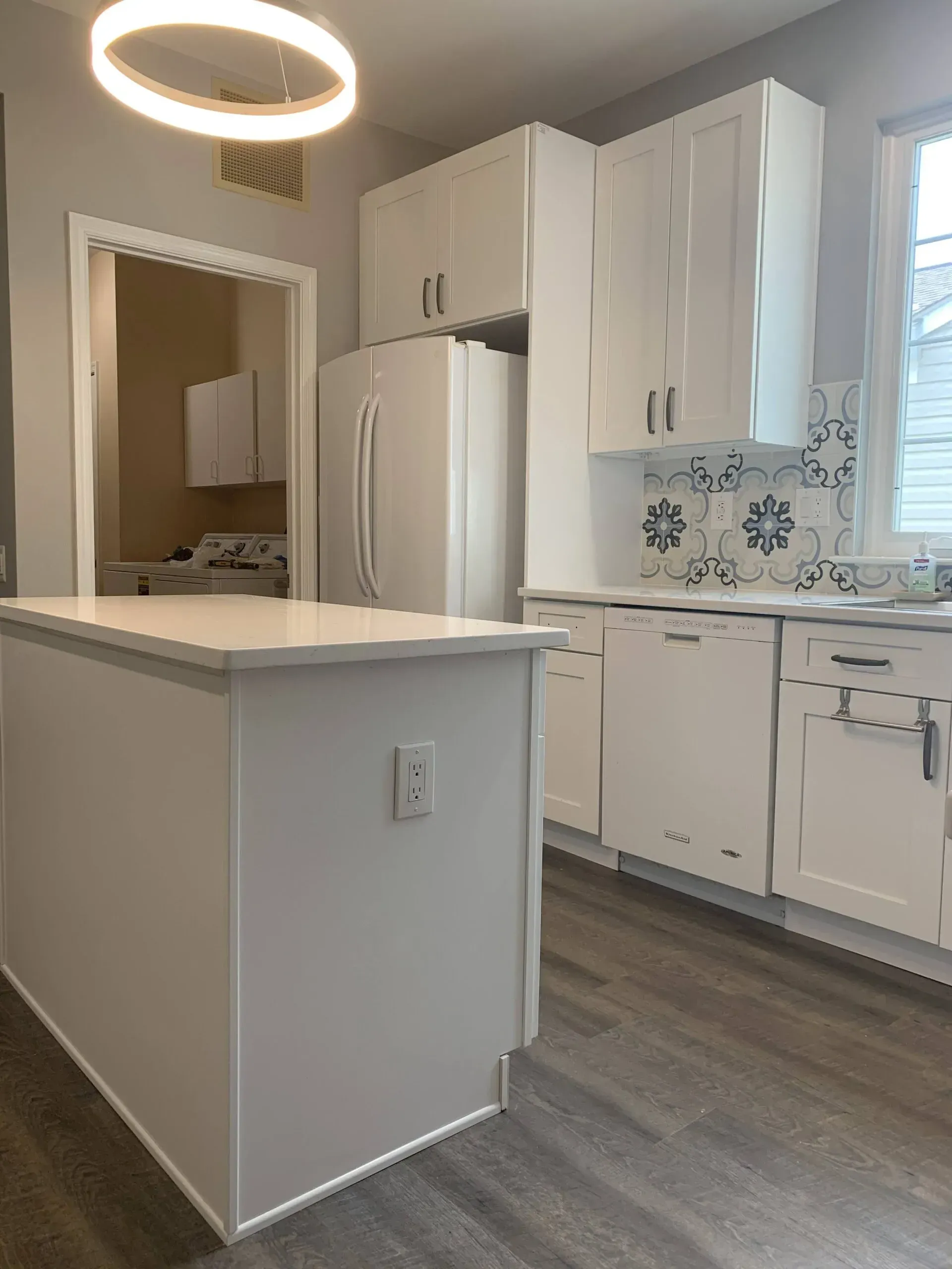 Bright white kitchen with island, cabinets, fridge, and snowflake tile backsplash, lit by a ring pendant light