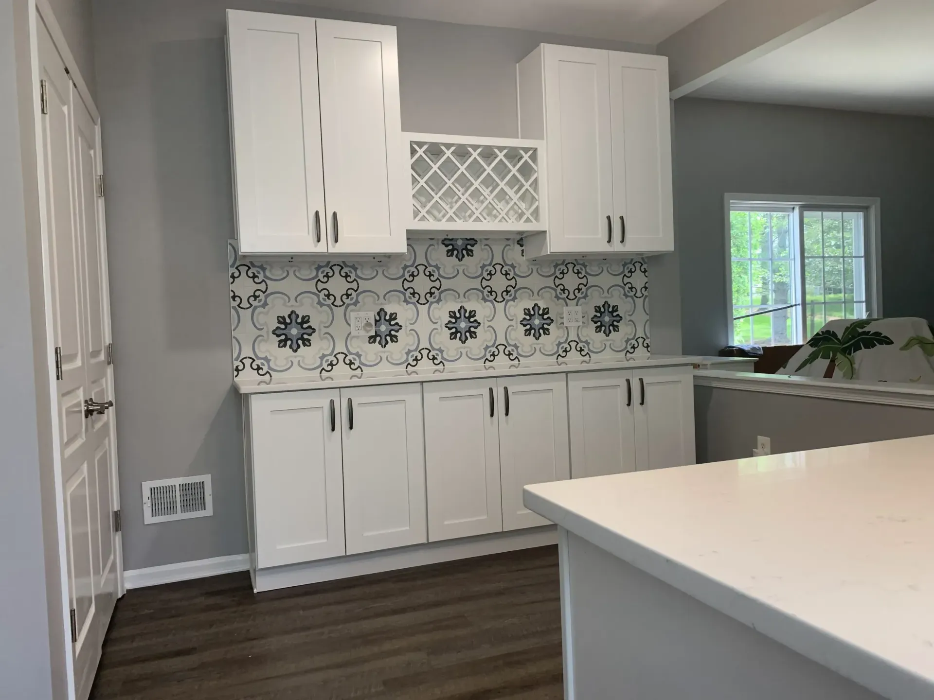 White kitchen with floral tile backsplash, white cabinets, and dark wood floors beside a bright window