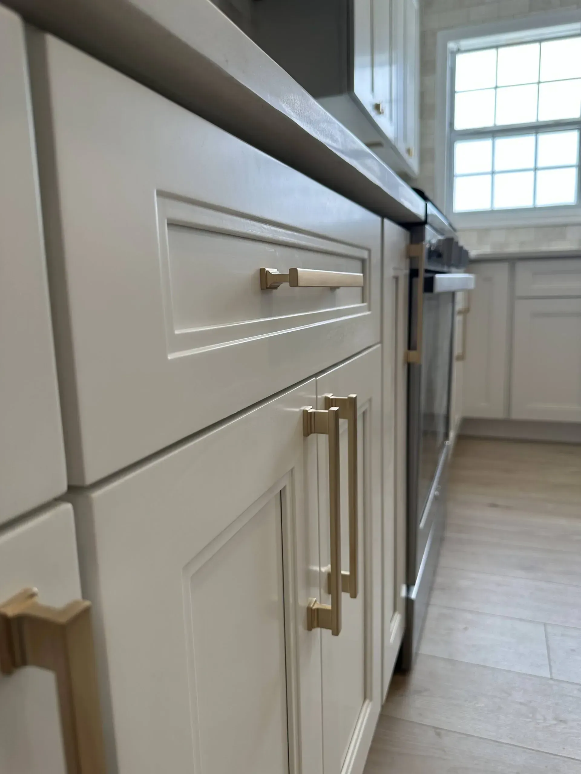 White kitchen cabinets with brass handles and a built-in stainless steel oven in a bright room.