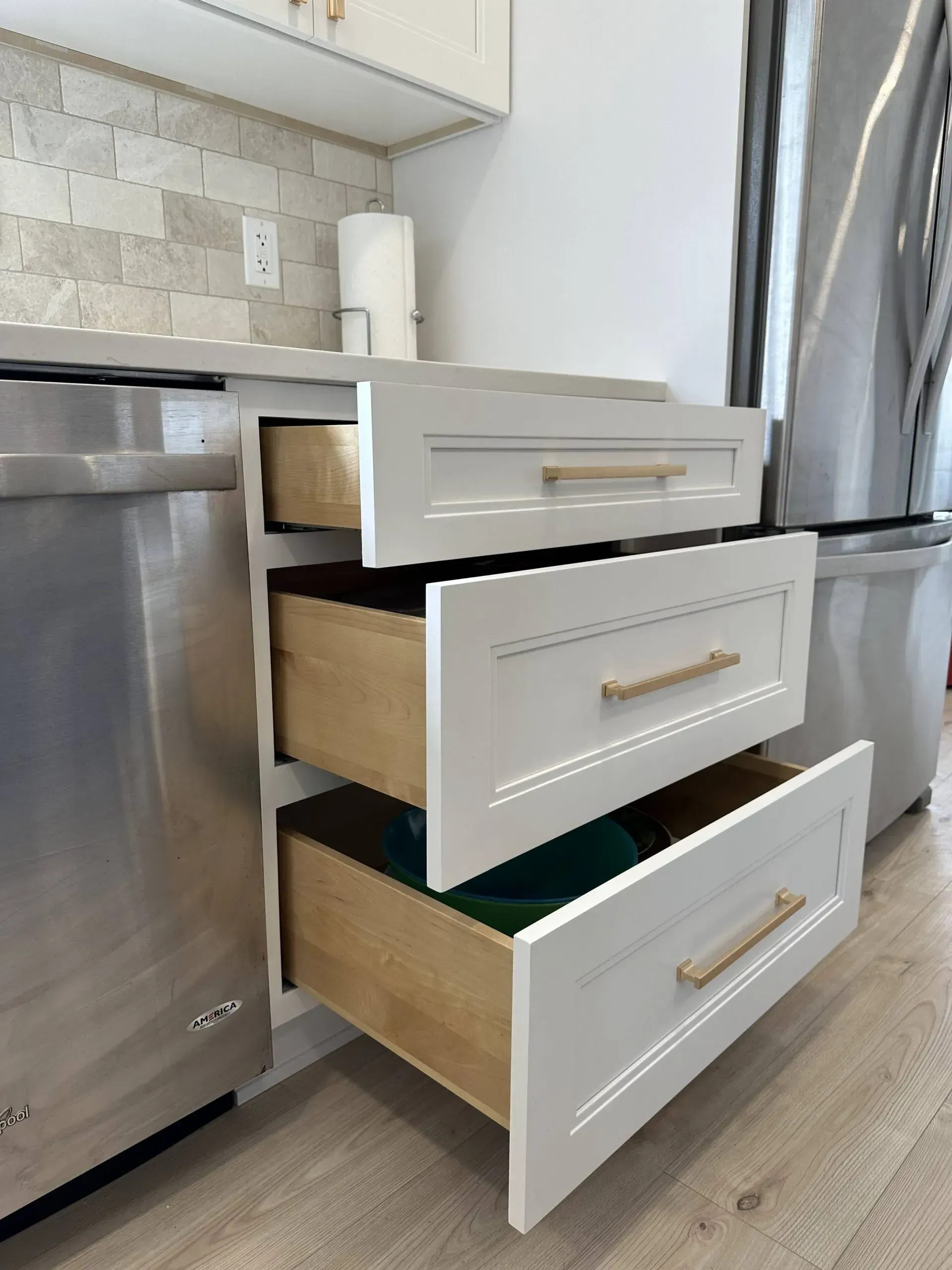 Three open drawers in a stainless steel kitchen cabinet beside a dishwasher and refrigerator.