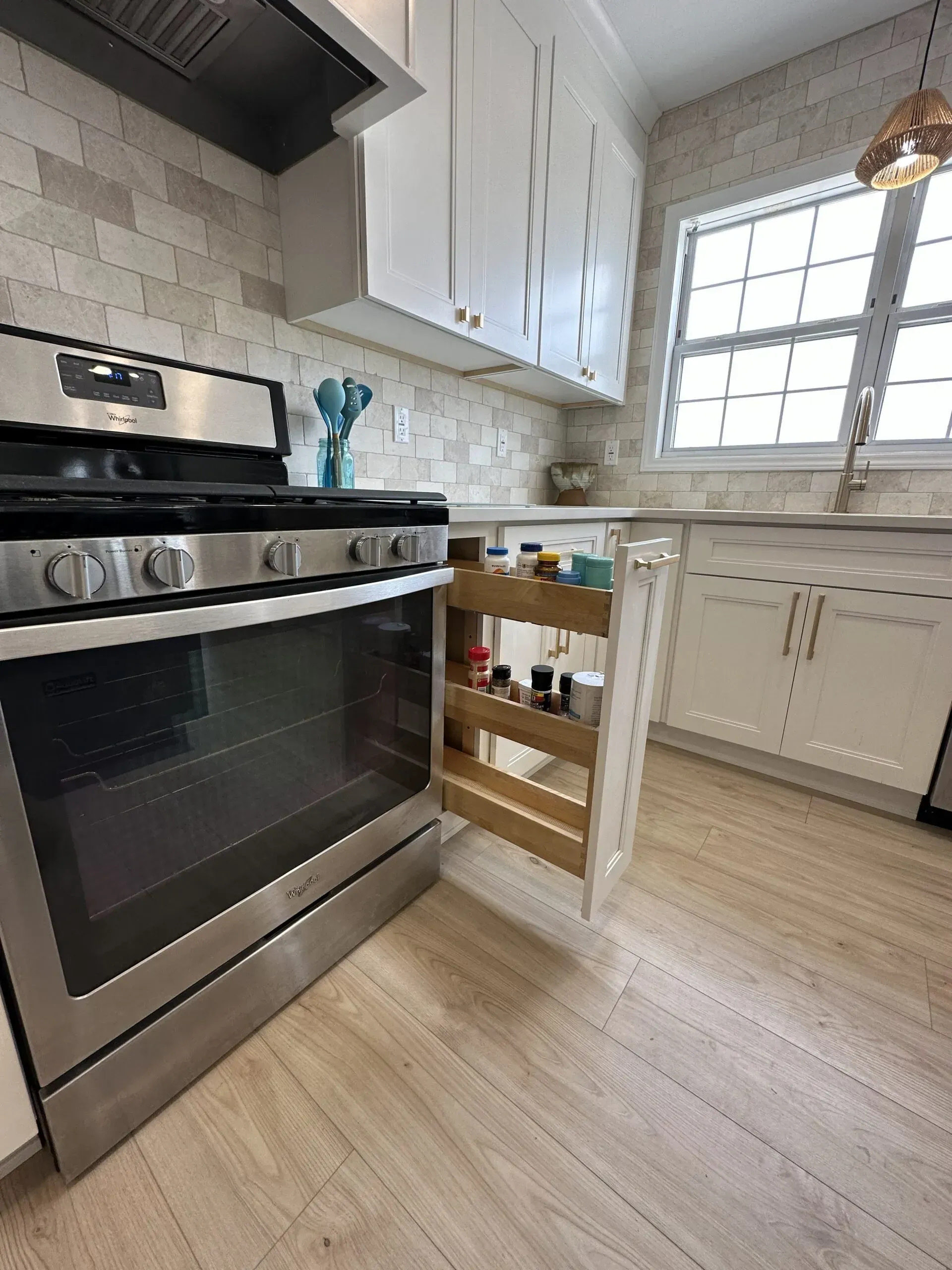 Bright kitchen with stainless steel stove, white cabinets, light wood floor, and a window over the sink.