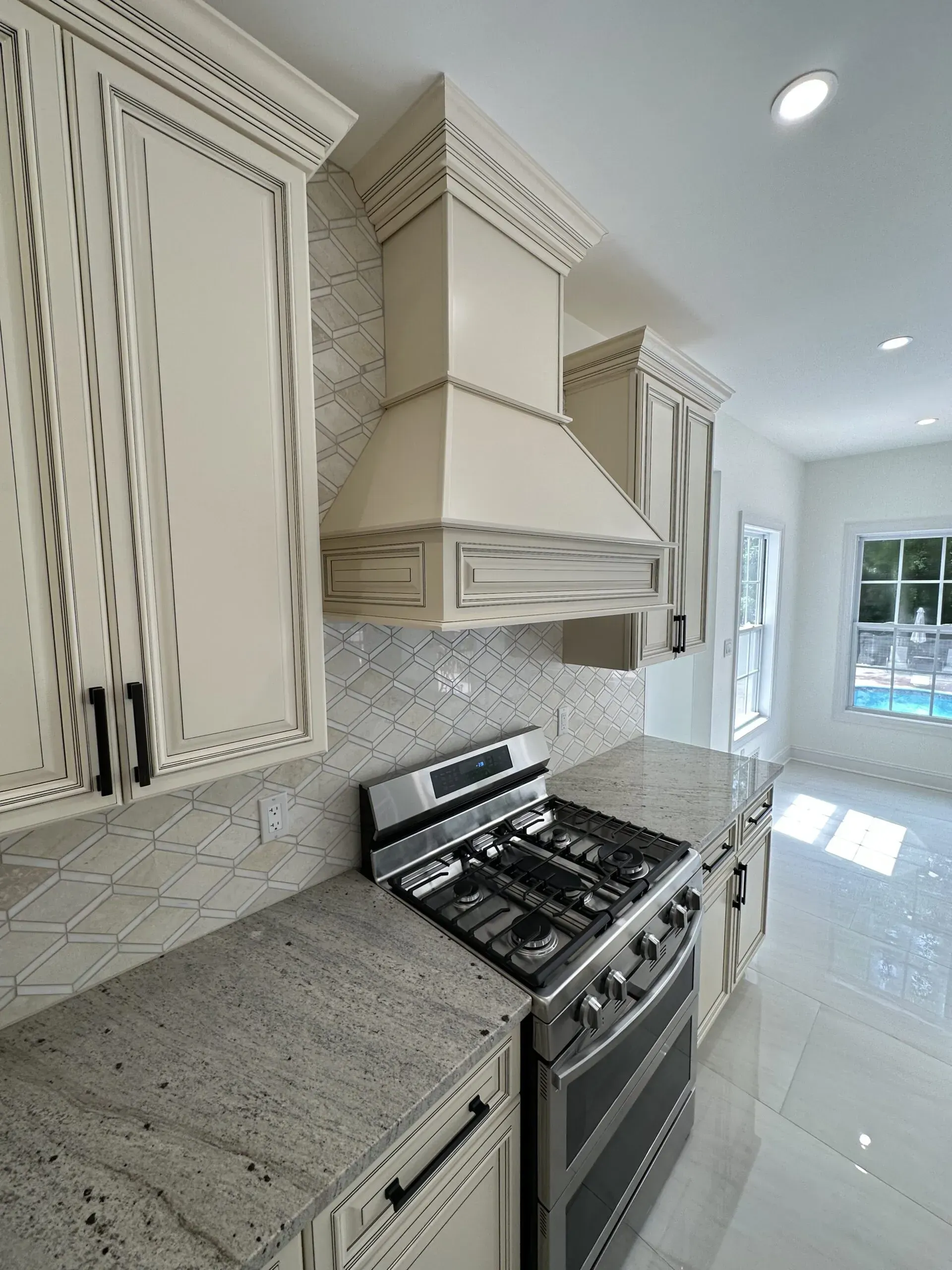 Bright white kitchen with granite counters, mosaic backsplash, and stainless gas range under a vent hood.