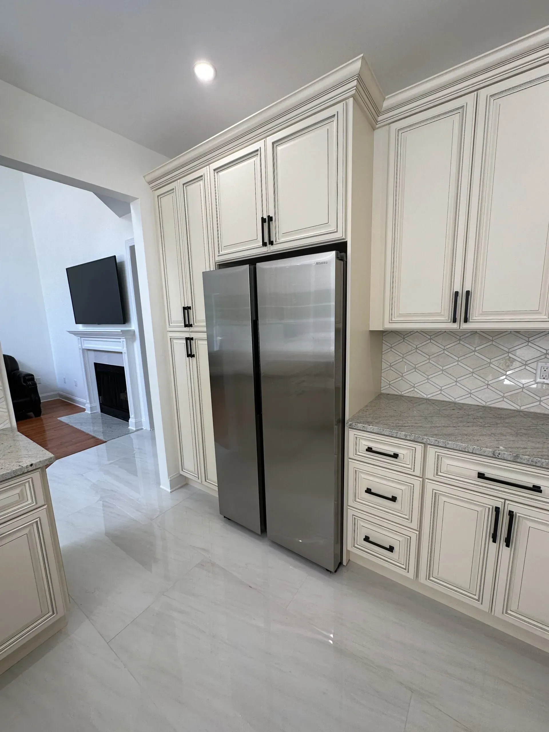 White kitchen with stainless steel refrigerator, cream cabinets, and tiled floor open to a living room.