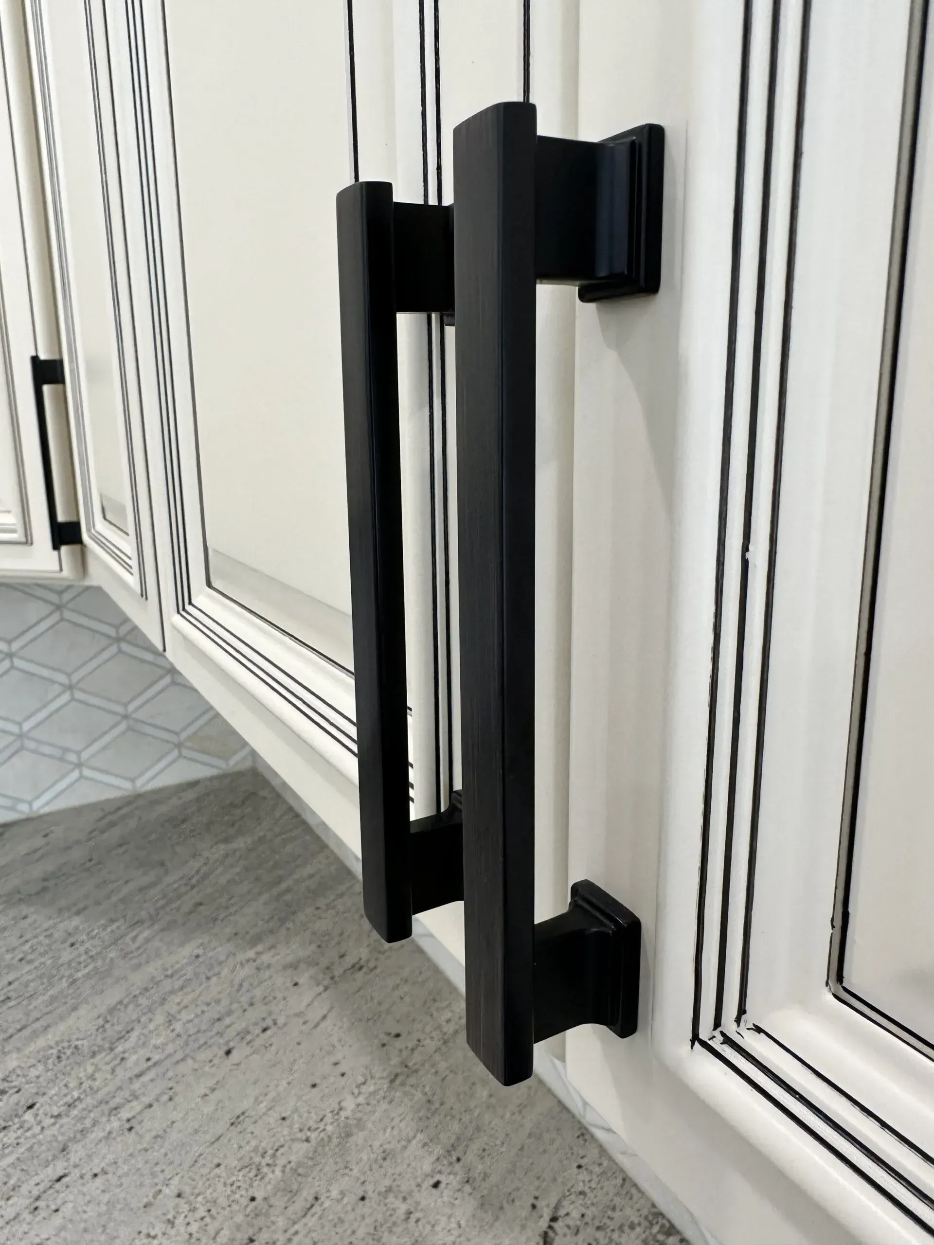 Black vertical handle on a white paneled cabinet door next to a gray floor.