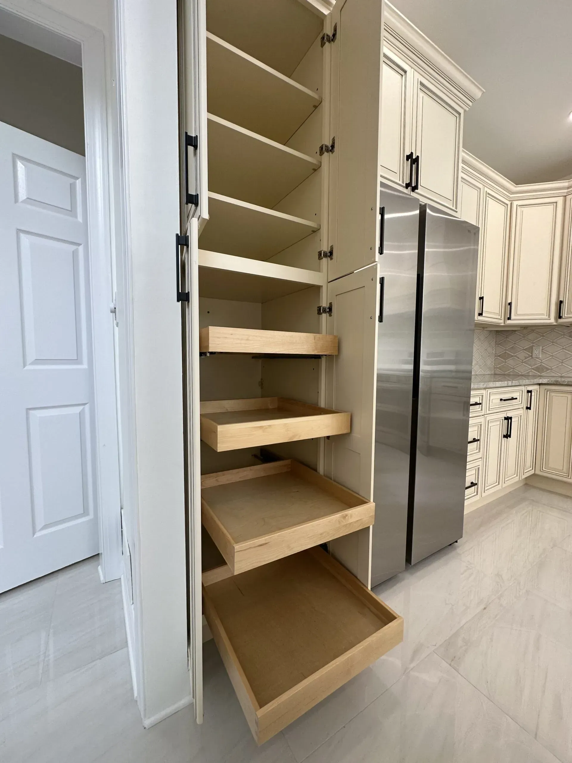 Built-in pantry cabinet with open wooden pull-out shelves next to stainless steel refrigerator in a bright kitchen