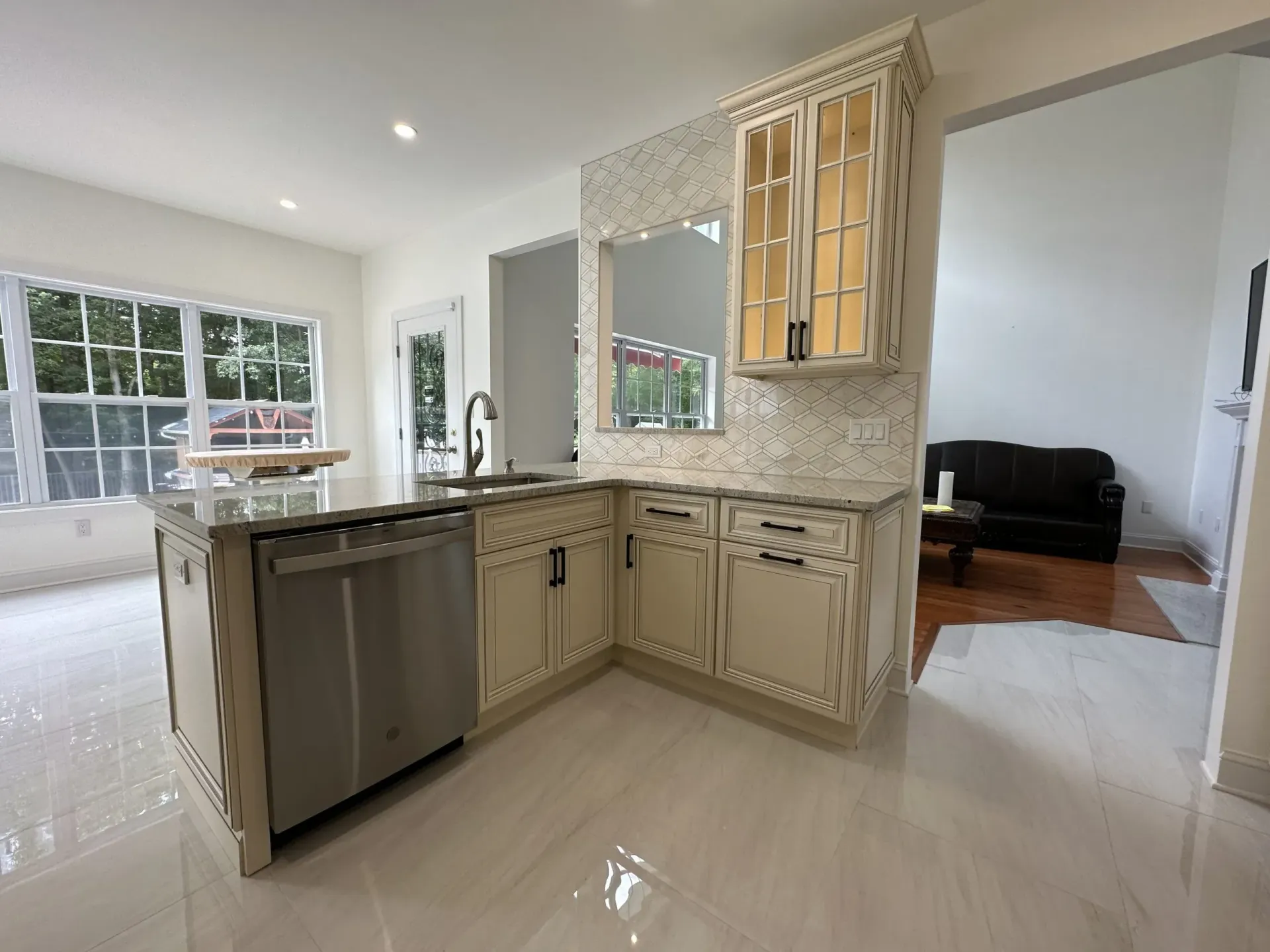 Bright kitchen with mosaic backsplash, beige cabinets, stainless dishwasher, and a view into a living room.