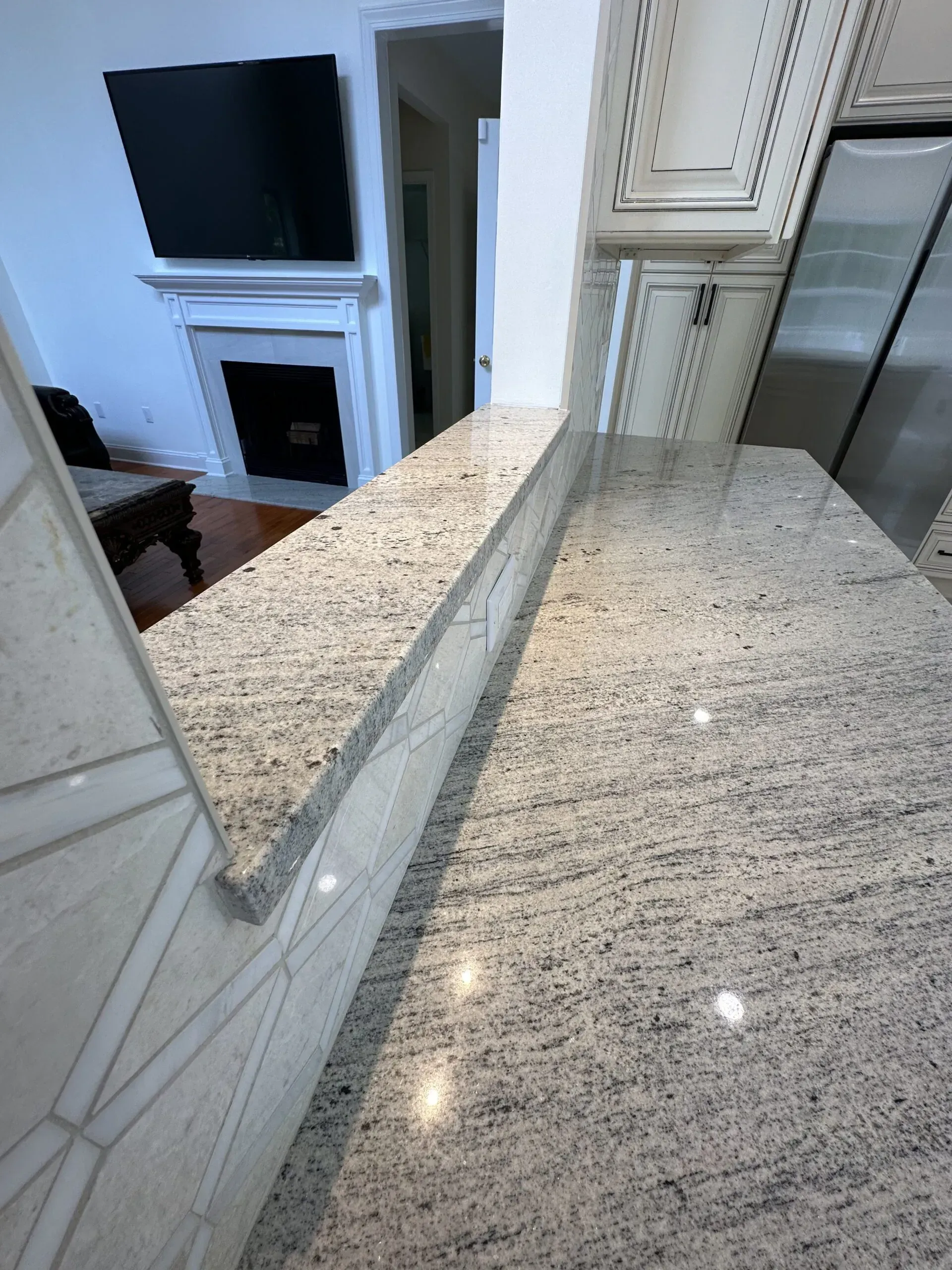Granite kitchen counter with mirrored backsplash and view into a living room with TV and fireplace