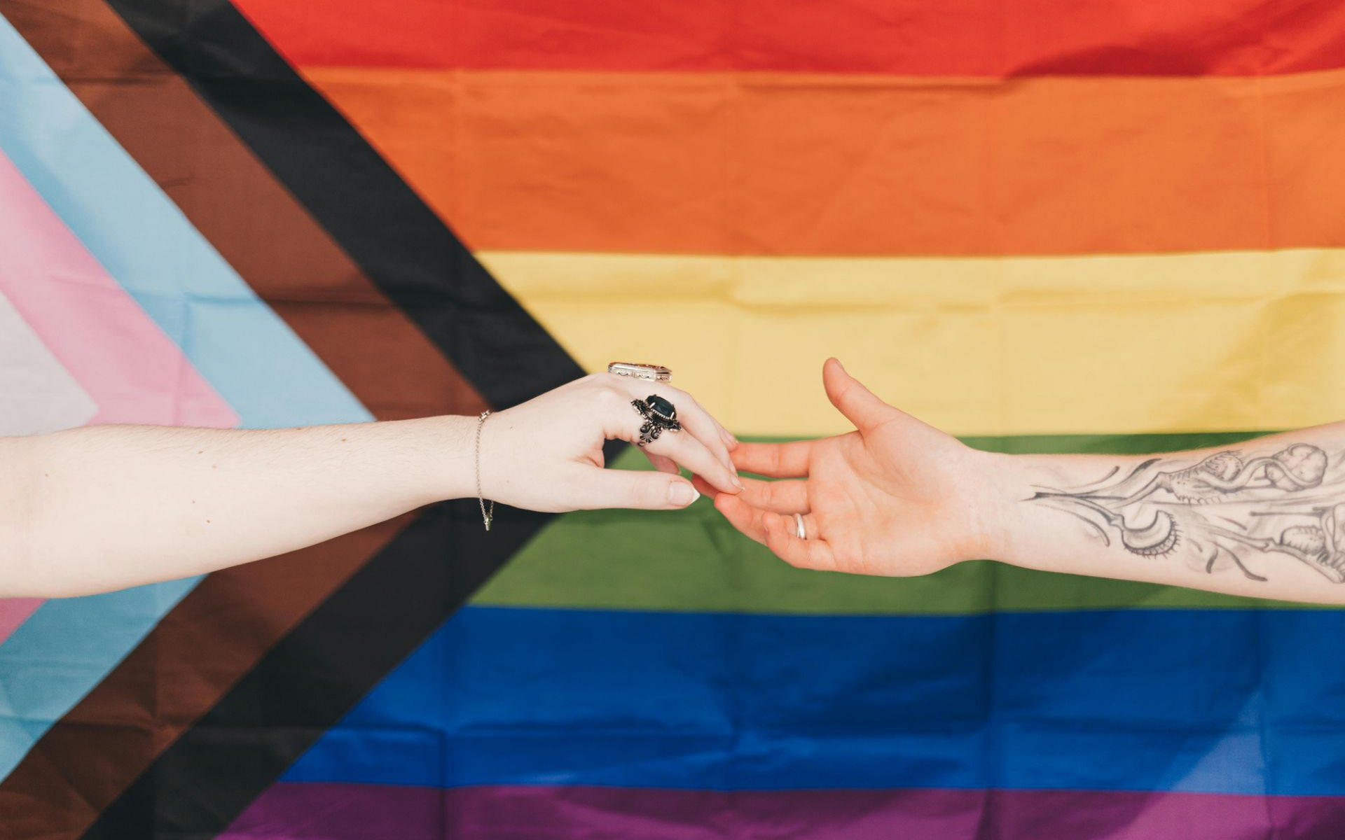 The hands of two people reach toward each other in front of a progress pride flag.