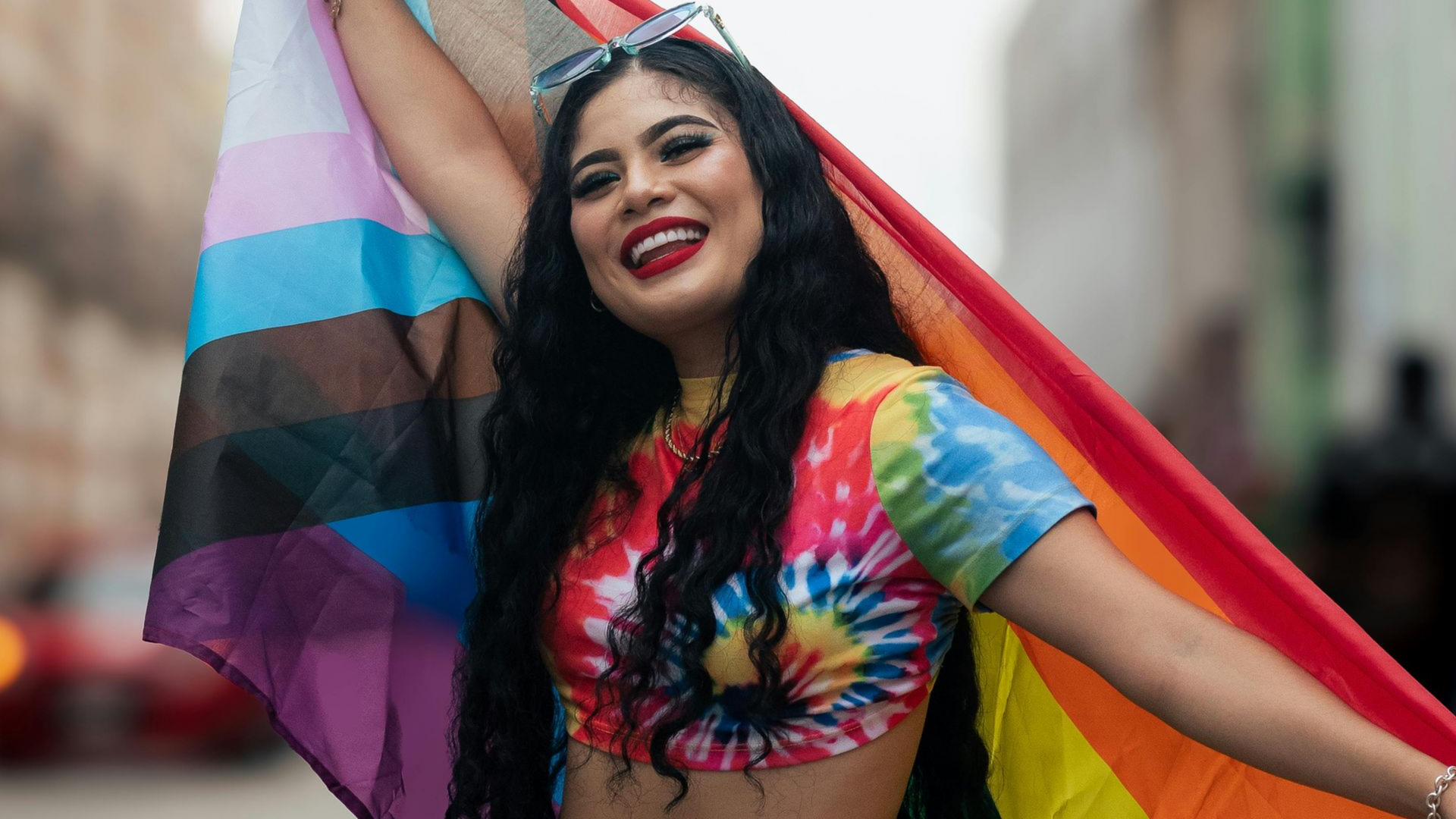 A person in a colorful tie-dye shirt smiles while holding a progress Pride flag behind them. 