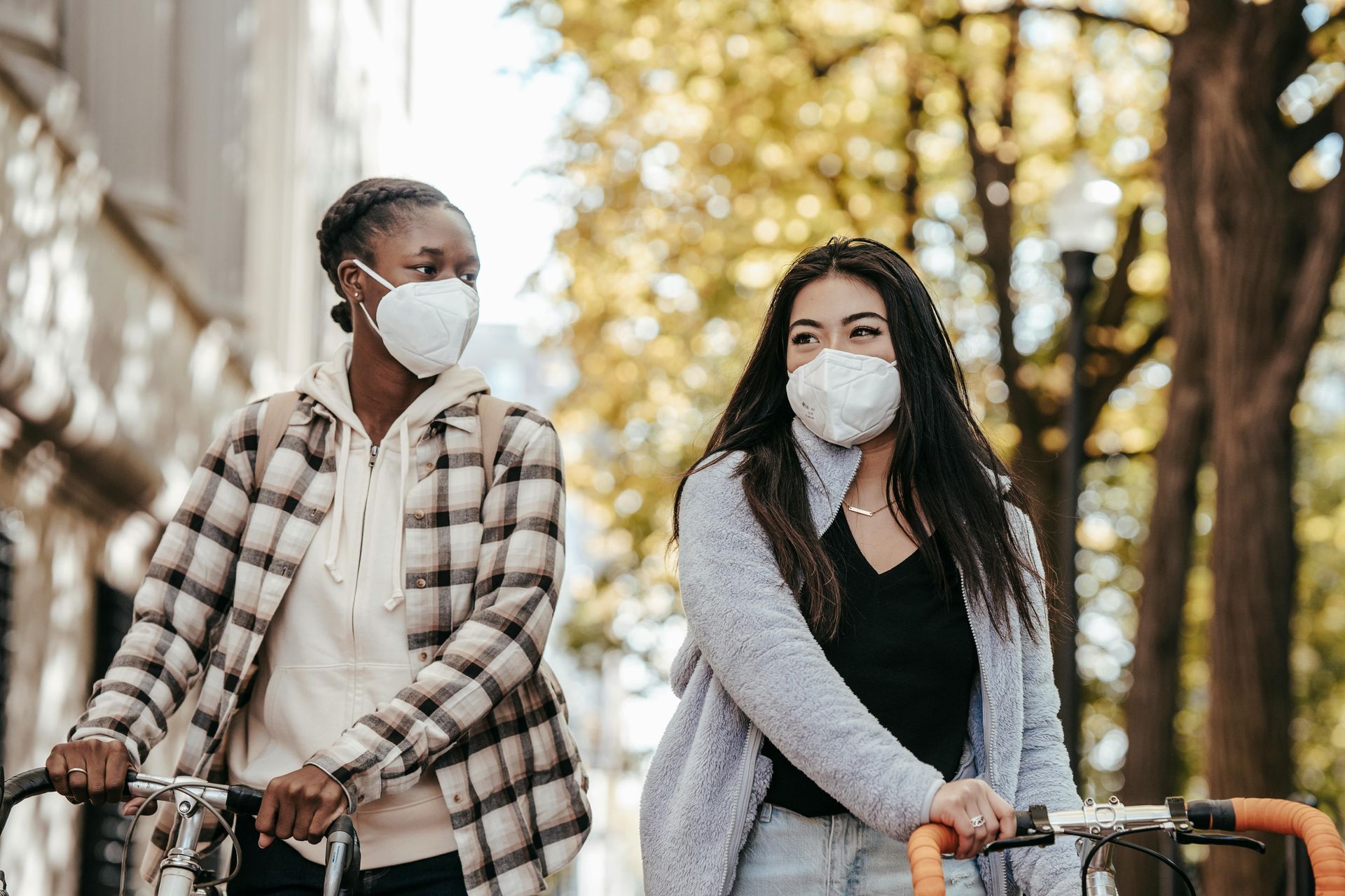 Two people wearing KN95 respirators walk their bicycles together down a sunny autumn street, appearing happy. Two people wearing KN95 respirators walk their bicycles together down a sunny autumn street, appearing happy.