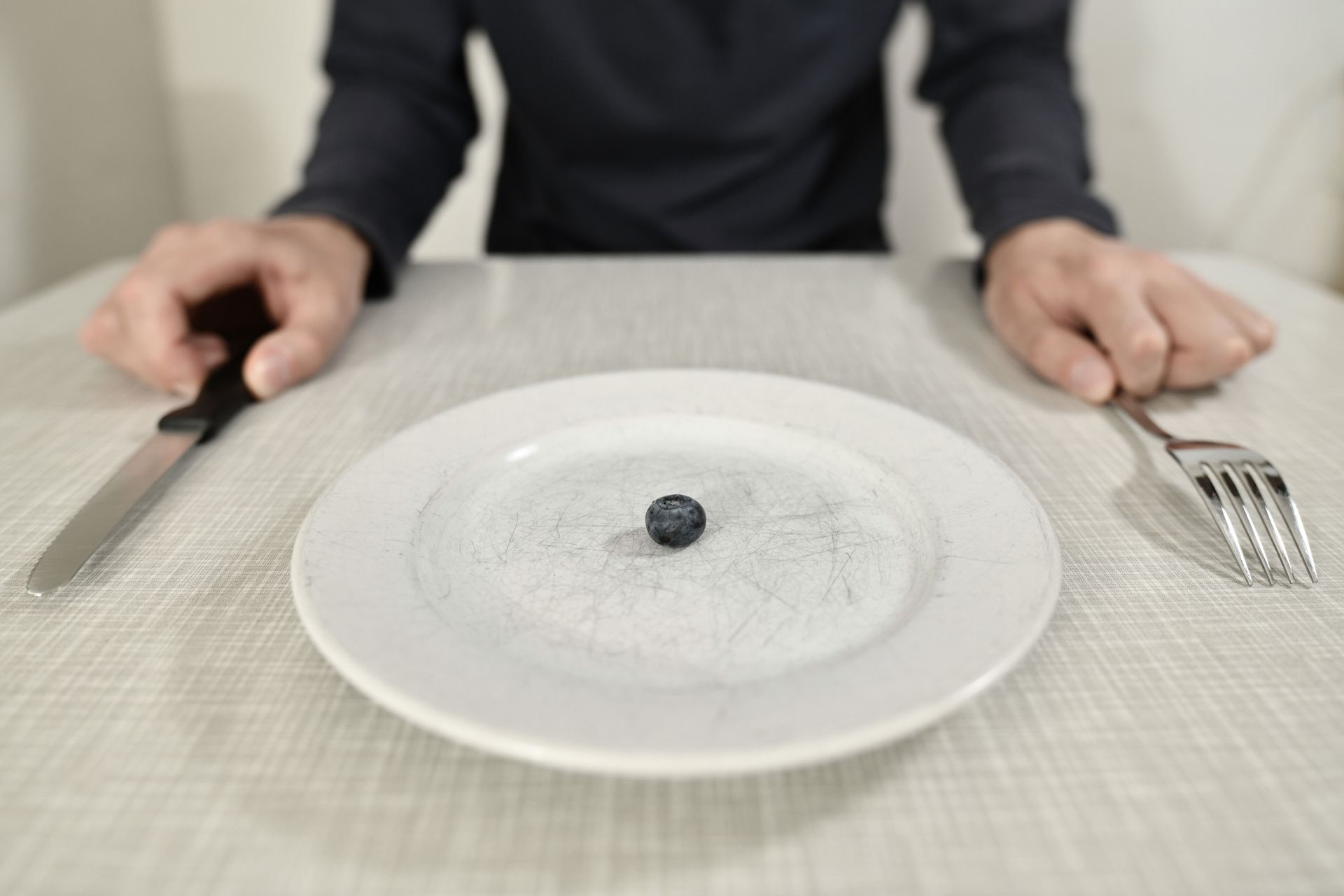 A person holding silverware sits in front of a plate holding only a small piece of food.