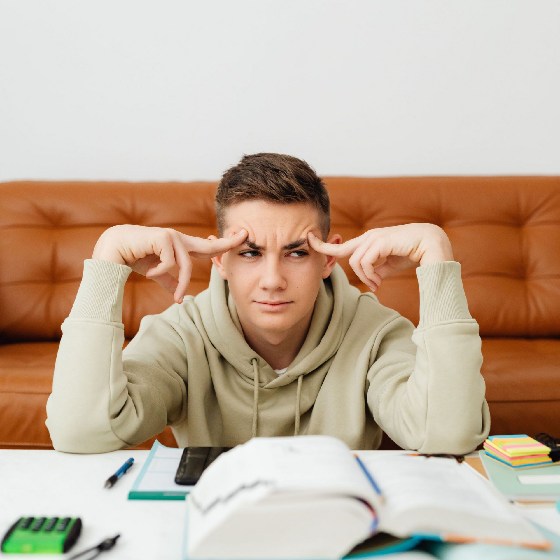 A young man sits at a desk in front of a pile of school supplies, looking frustrated.