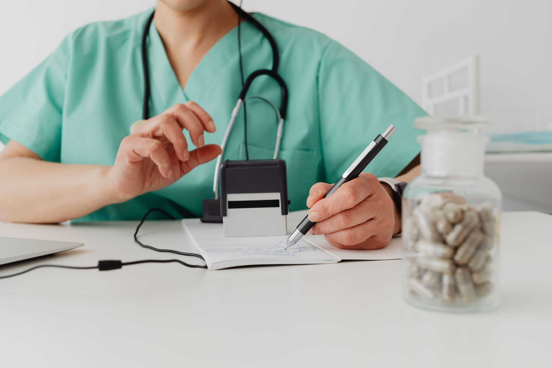 A physician in blue scrubs sits at a desk, writing on a prescription pad.